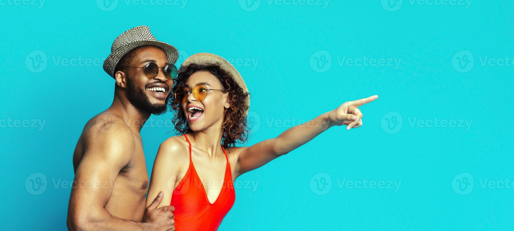 Black couple standing in front of a bright blue background, both wearing summer clothes. The woman is pointing to the right with a smile on her face, and the man is looking at her with a loving smile photo