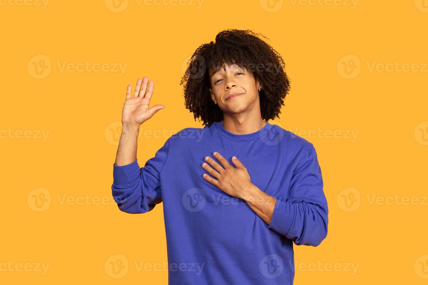 A young man with curly black hair, wearing a blue sweatshirt, is standing against a yellow background. He is waving with his right hand and placing his left hand over his chest, smiling gently. photo
