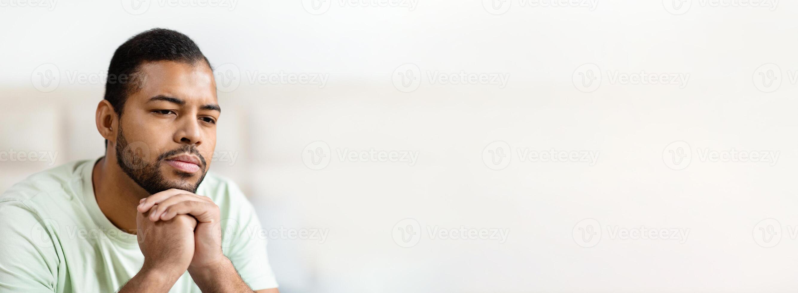 African American man sits quietly in a light green shirt, deep in thought with his hands clasped. The soft lighting creates a calm atmosphere, suggesting a moment of introspection and contemplation. photo
