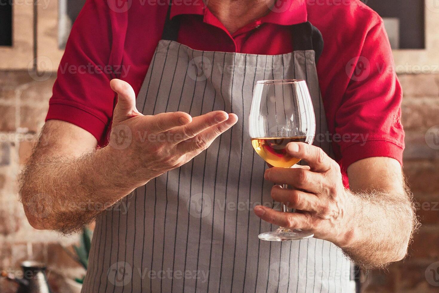 A senior man, wearing glasses and an apron, stands in a modern kitchen with a brick wall. He is holding a glass of wine and smiling, enjoying his time while preparing a meal, cropped photo