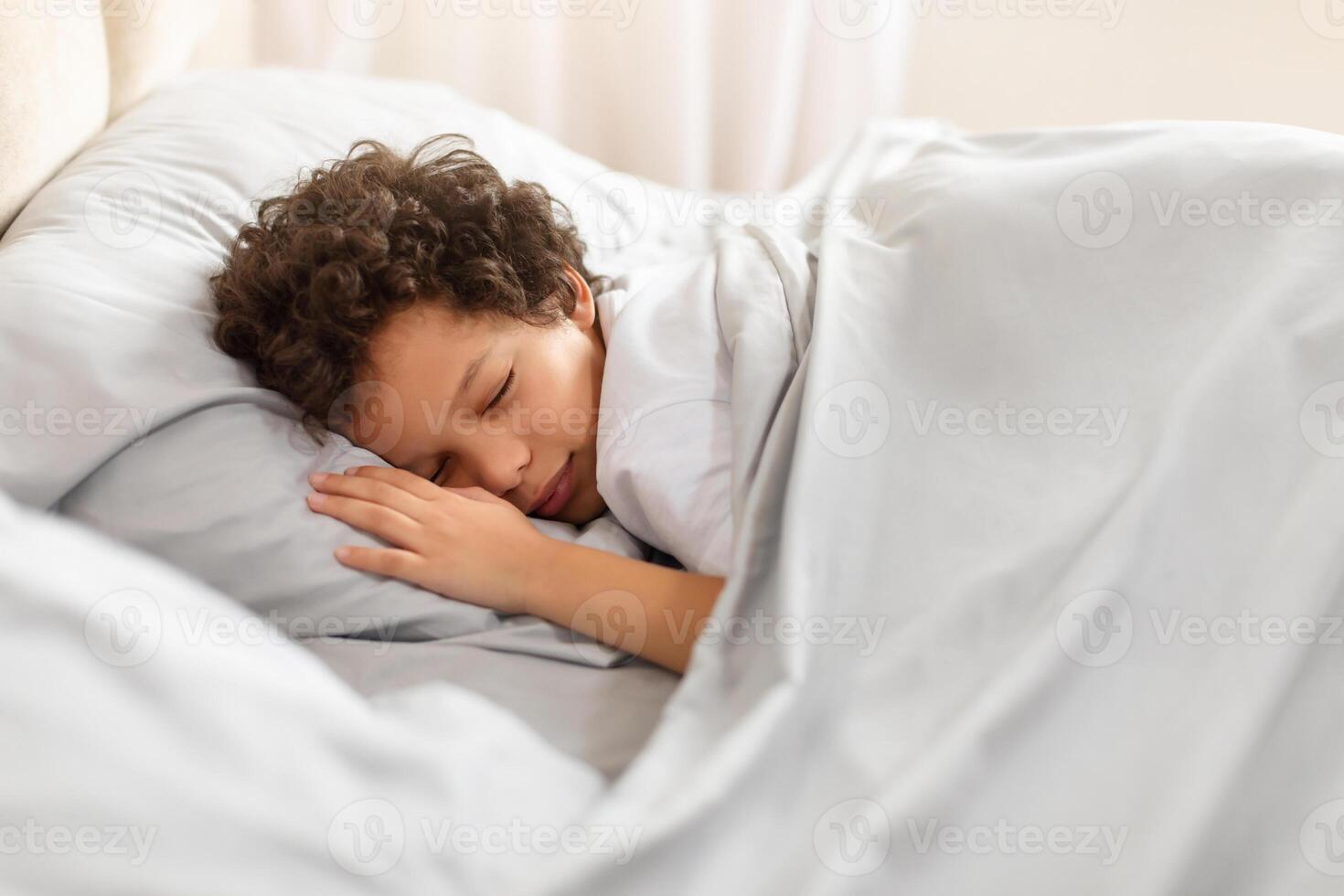 A young African American boy is peacefully sleeping in a bed with crisp white sheets covering him. His head rests on a fluffy pillow as he finds rest and comfort in his surroundings. photo