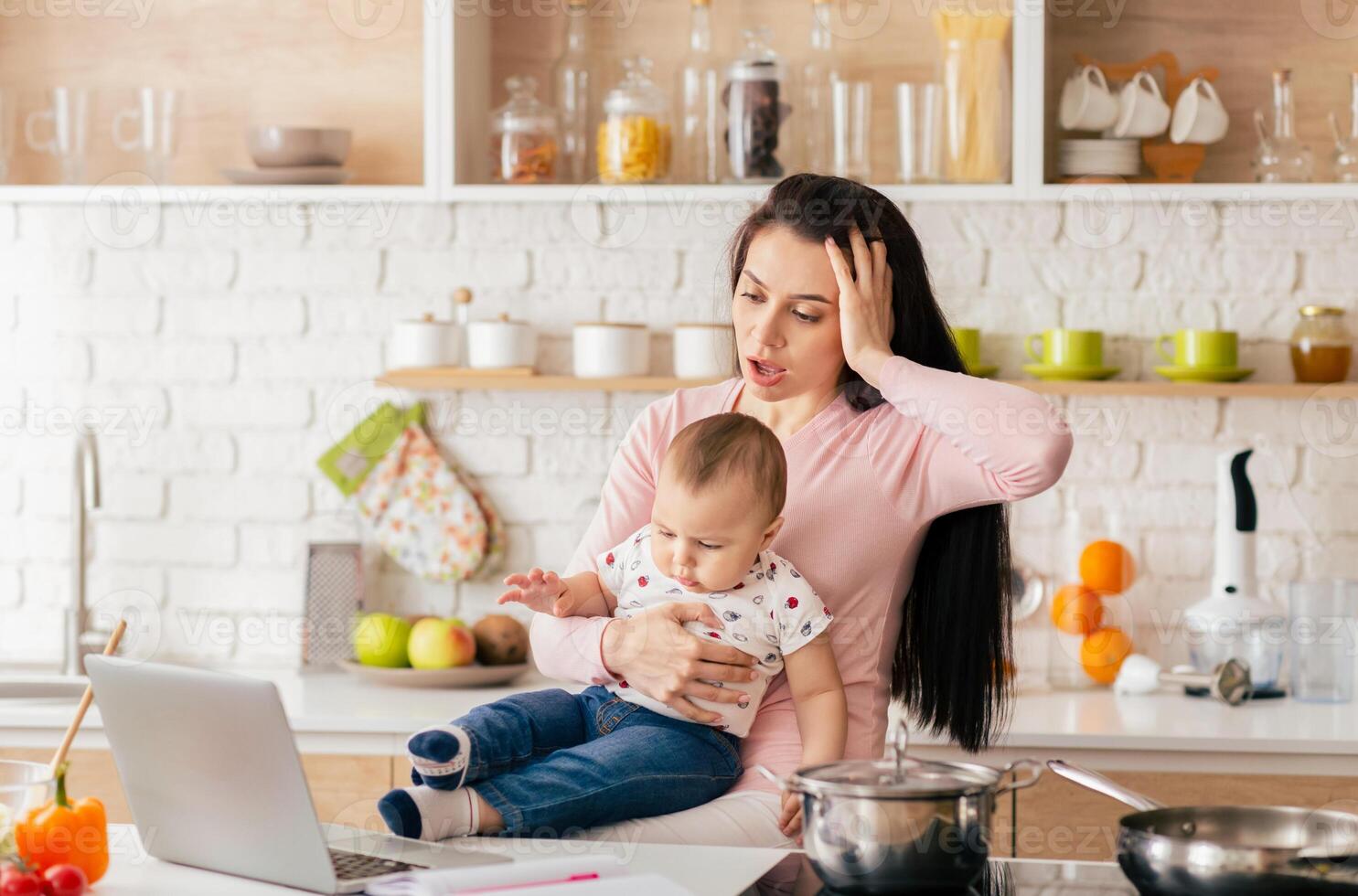 A mother tries to manage her remote work on a laptop while holding her baby in a modern kitchen, highlighting the challenges of multitasking with household duties and childcare responsibilities. photo
