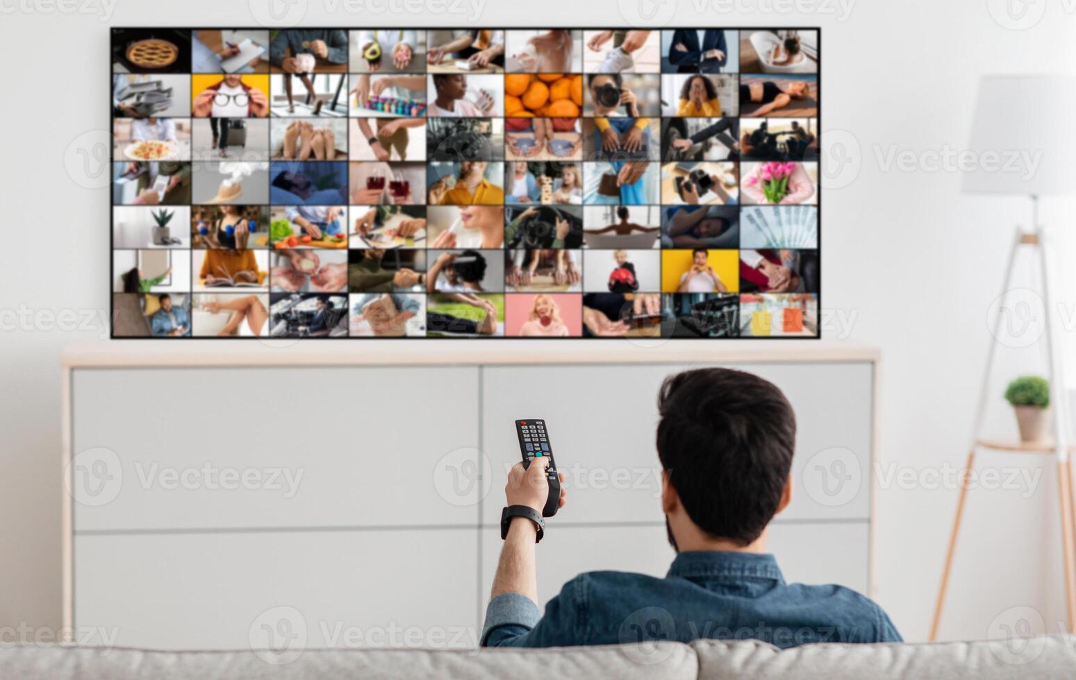 A man is sitting on a couch and watching television, holding the remote control in his hand. The TV screen is showing a grid of images, each depicting a different scene. photo