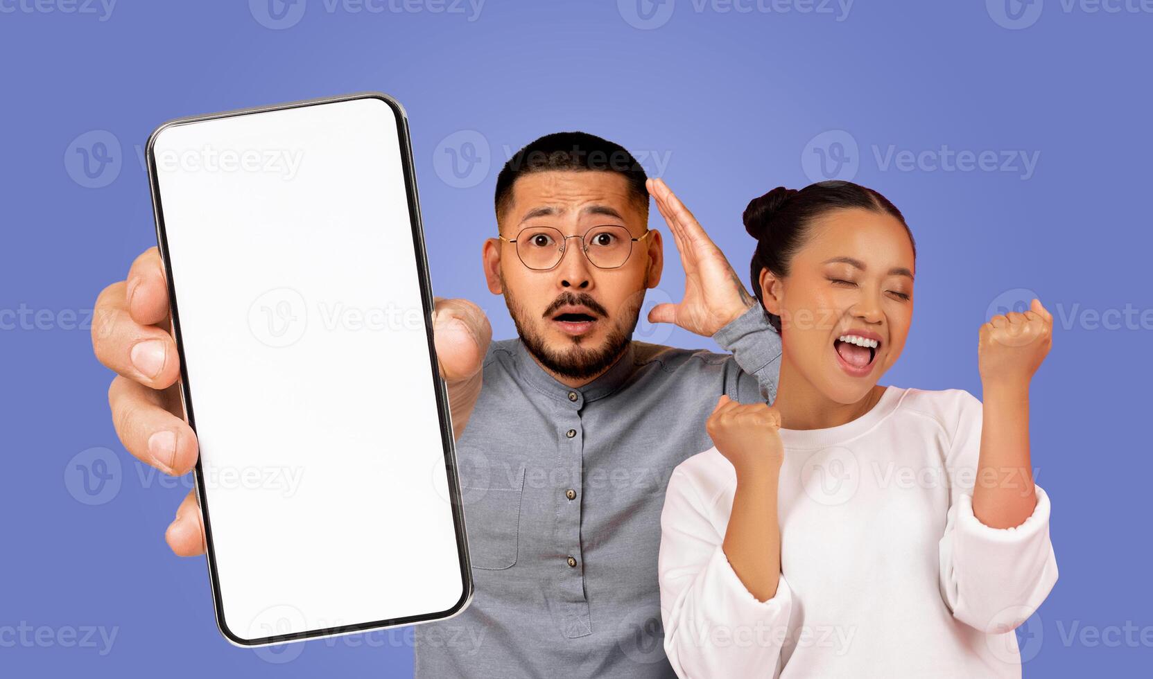Asian man and woman stand against a purple backdrop. The man holds a smartphone with a blank screen in front of him while he looks surprised photo