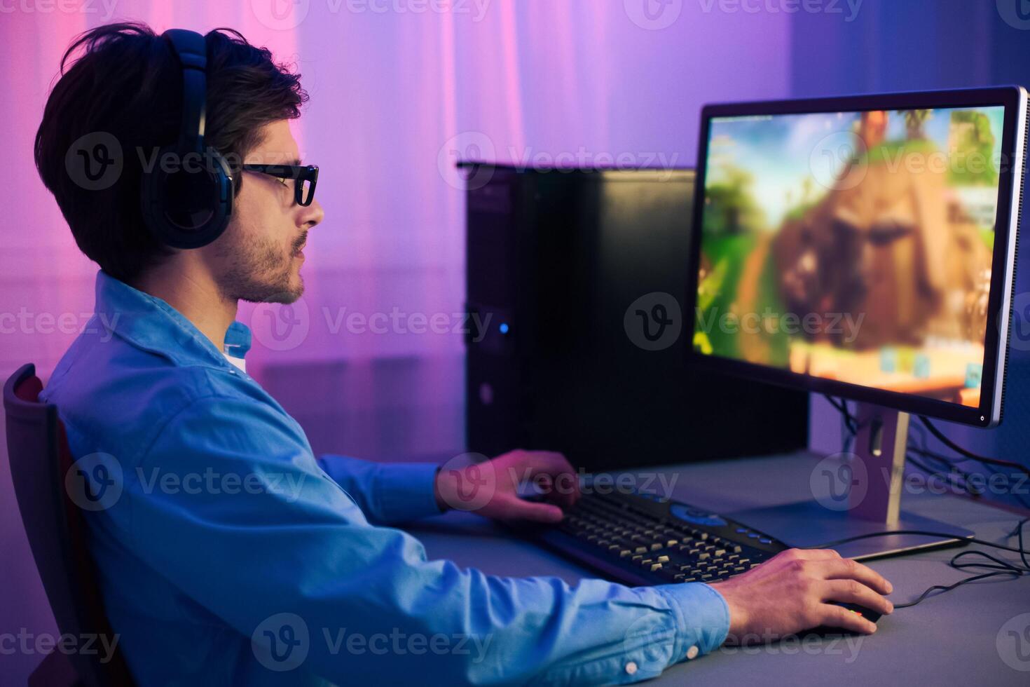 A young man wearing headphones is playing a computer game at a desk in his home office. He is focused on the screen and is using a keyboard and mouse to control the game. photo