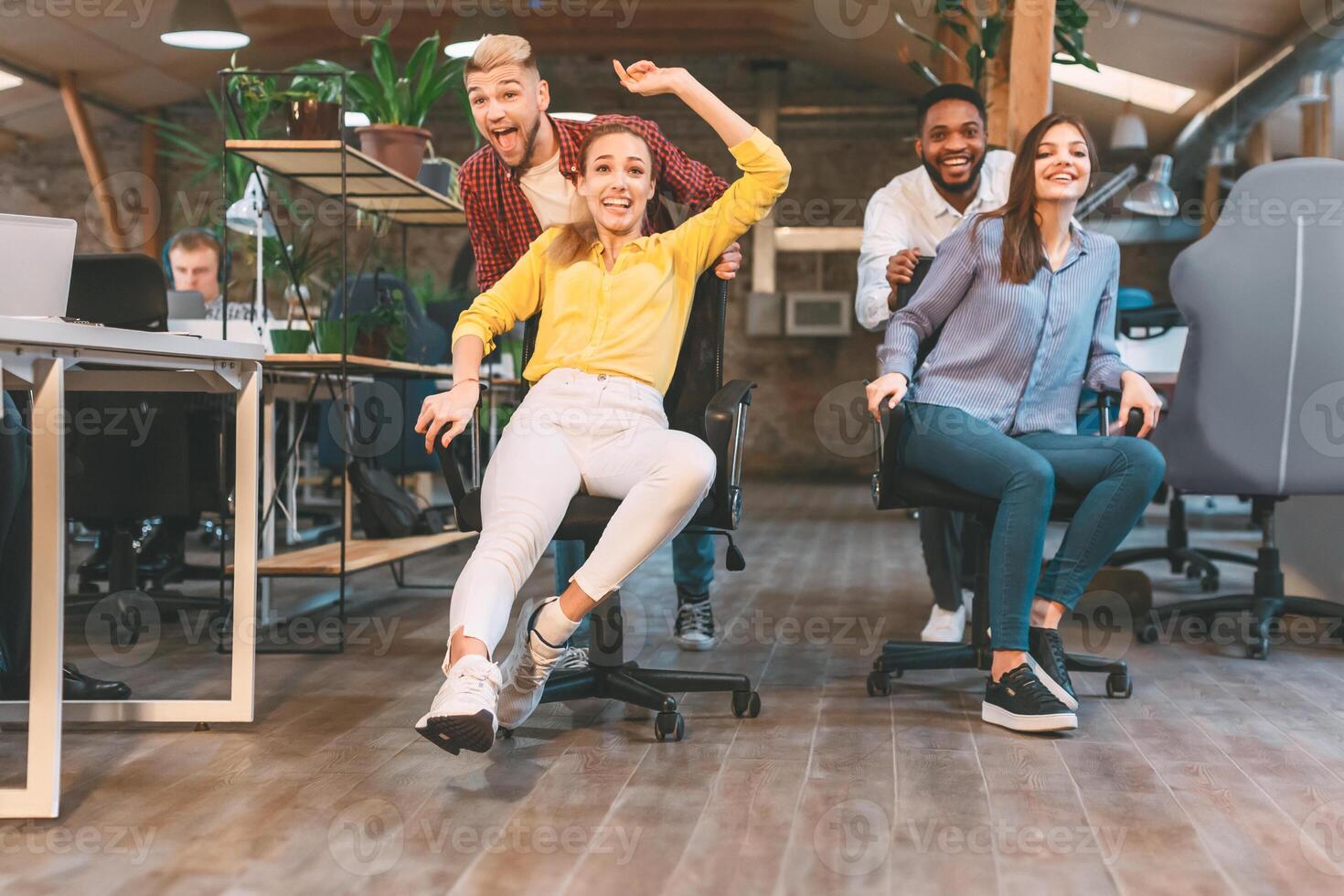 Time for break. Joyful colleagues having fun at workplace, making office chair race in coworking space photo