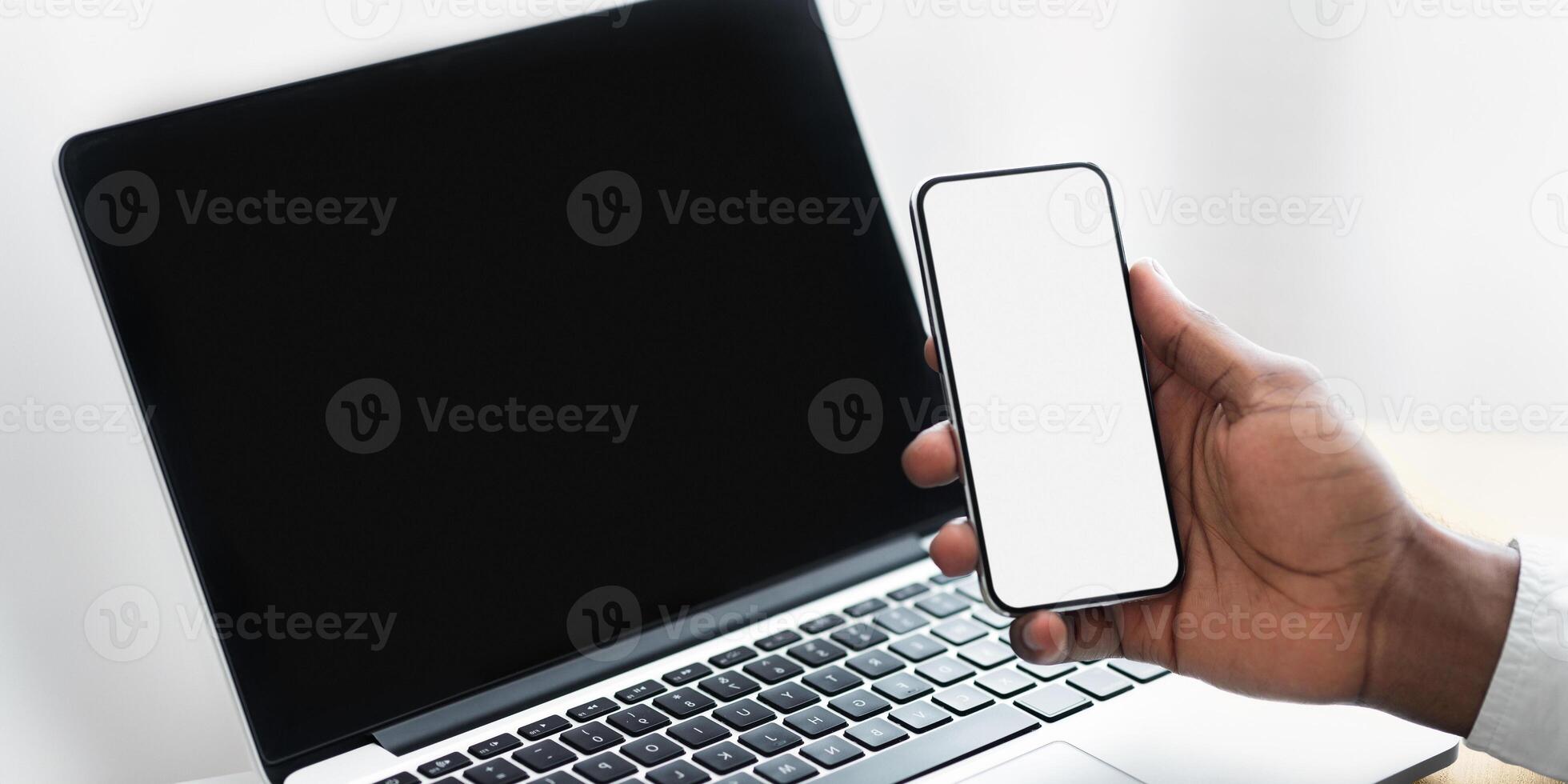 A hand holds a smartphone with a blank screen near a laptop with a black screen. photo