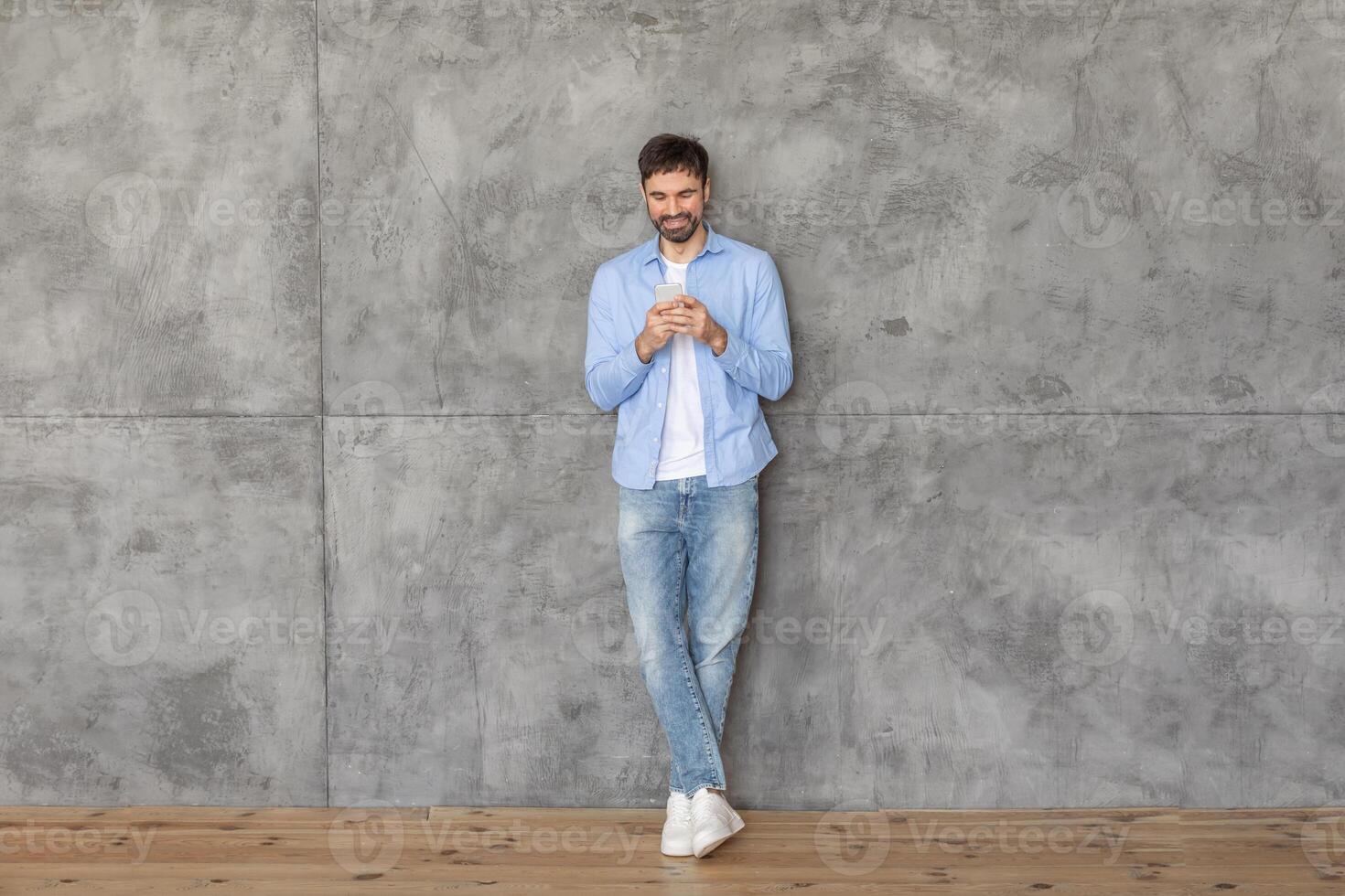 A young man in a blue shirt and jeans stands with his back against a concrete wall. He is using his smartphone and smiling as he looks down at the screen. The floor is made of wood. photo