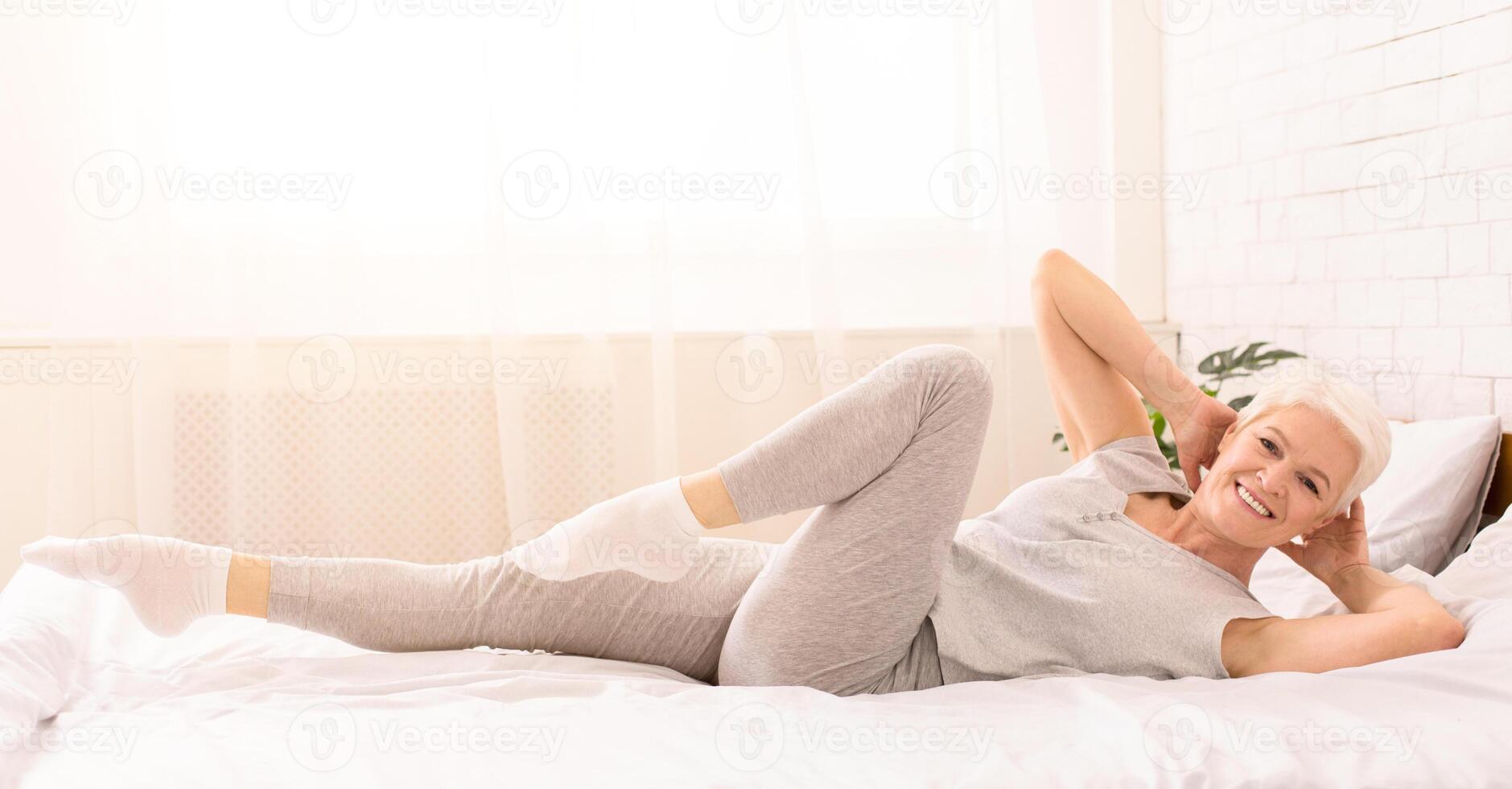 A cheerful senior woman is laying on a white bed, stretching her body and enjoying a morning exercise routine in a brightly lit and serene bedroom environment. photo
