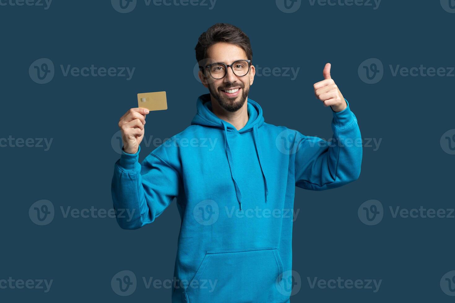 A young man in a blue hoodie smiles and gives a thumbs-up while holding a card. The background is a solid color, emphasizing his cheerful expression and positive energy. photo