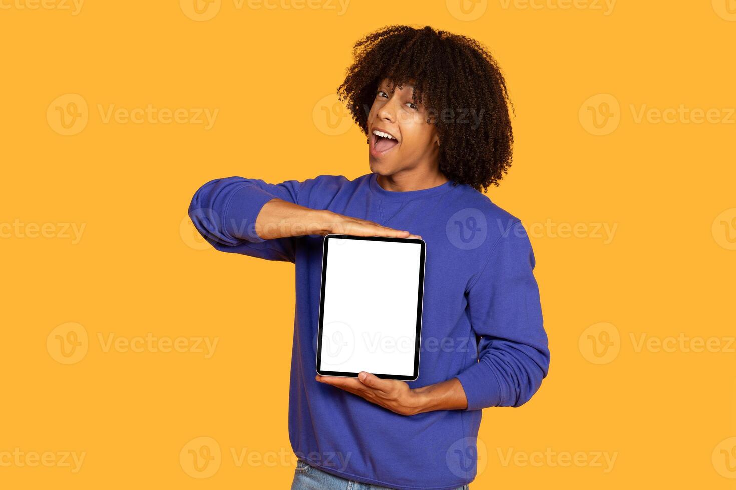 A young man with curly hair is standing against a yellow background. He is wearing a blue sweatshirt and is holding a digital tablet in front of him. The tablet has a blank screen. photo