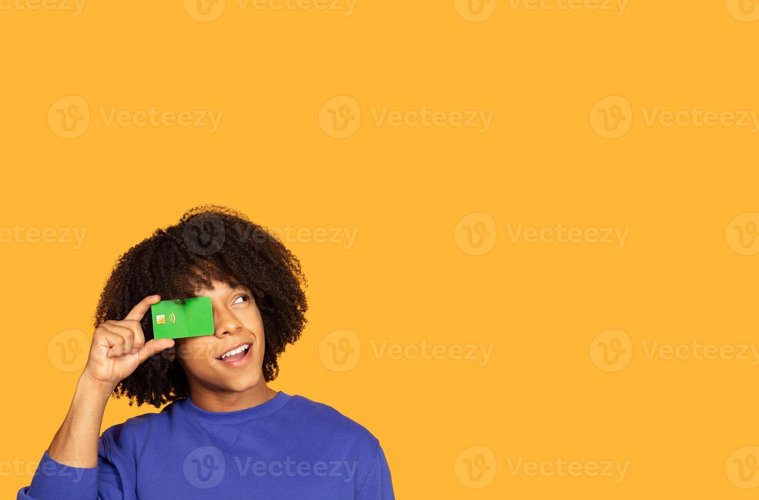 A young man with curly hair holds a green card near his face, gazing upwards with an expression of curiosity and delight. The bright yellow background adds a cheerful tone to the moment, copy space photo