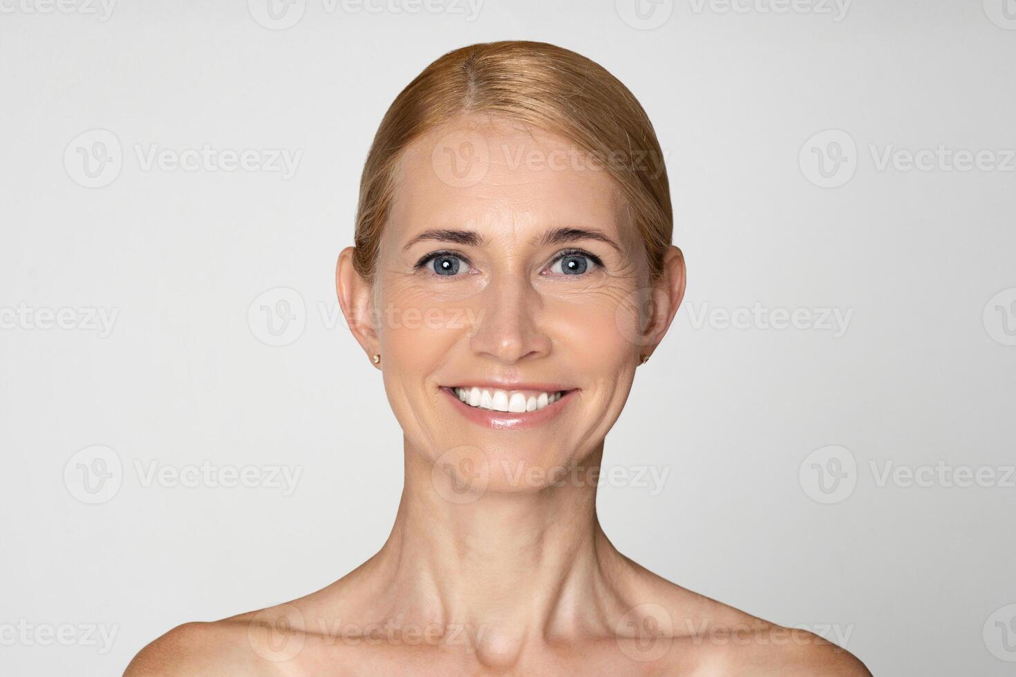 This photo shows a close-up portrait of a middle-aged woman with blonde hair, pulled back from her face, and blue eyes. She has a bright, genuine smile, revealing her white teeth.