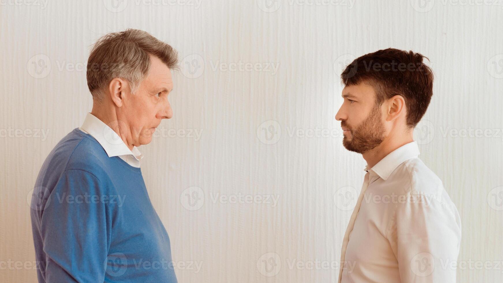Conflict Between Generations. Elderly Man And Mature Son Looking Severely At Each Other Standing Face-To-Face Against White Wall Indoor. Panorama photo