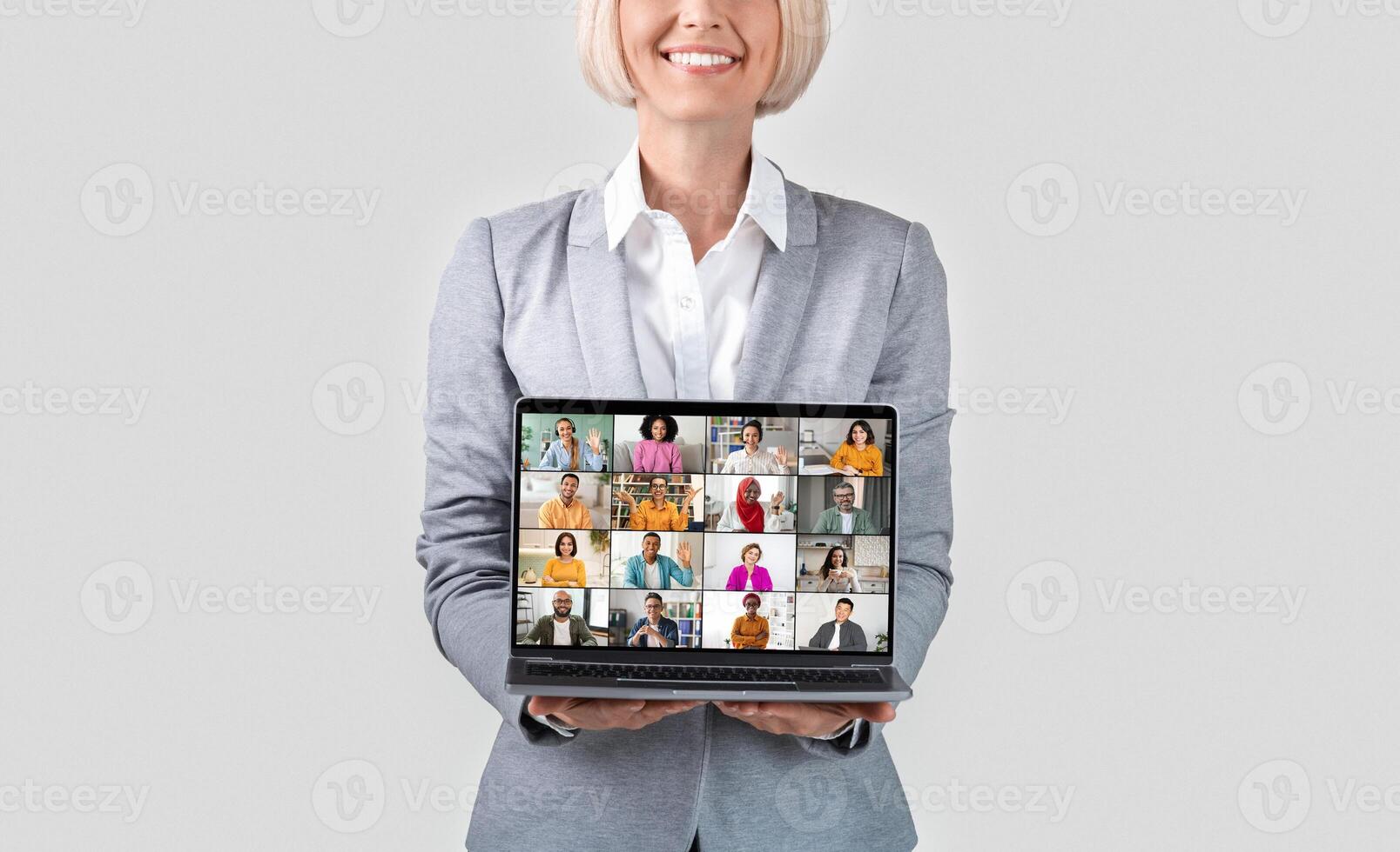 A smiling woman with blonde hair is holding a laptop in front of her. The laptop screen displays a conference with 16 participants, all diverse individuals with varying ethnicities and genders photo