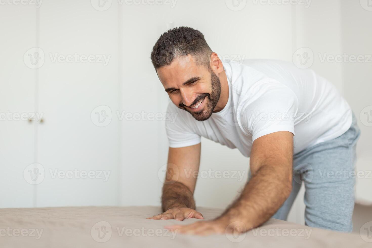 A man in a white t-shirt and grey sweatpants is smiling as he makes his bed in a bedroom. His arms are outstretched, and he is smoothing the sheets down. photo