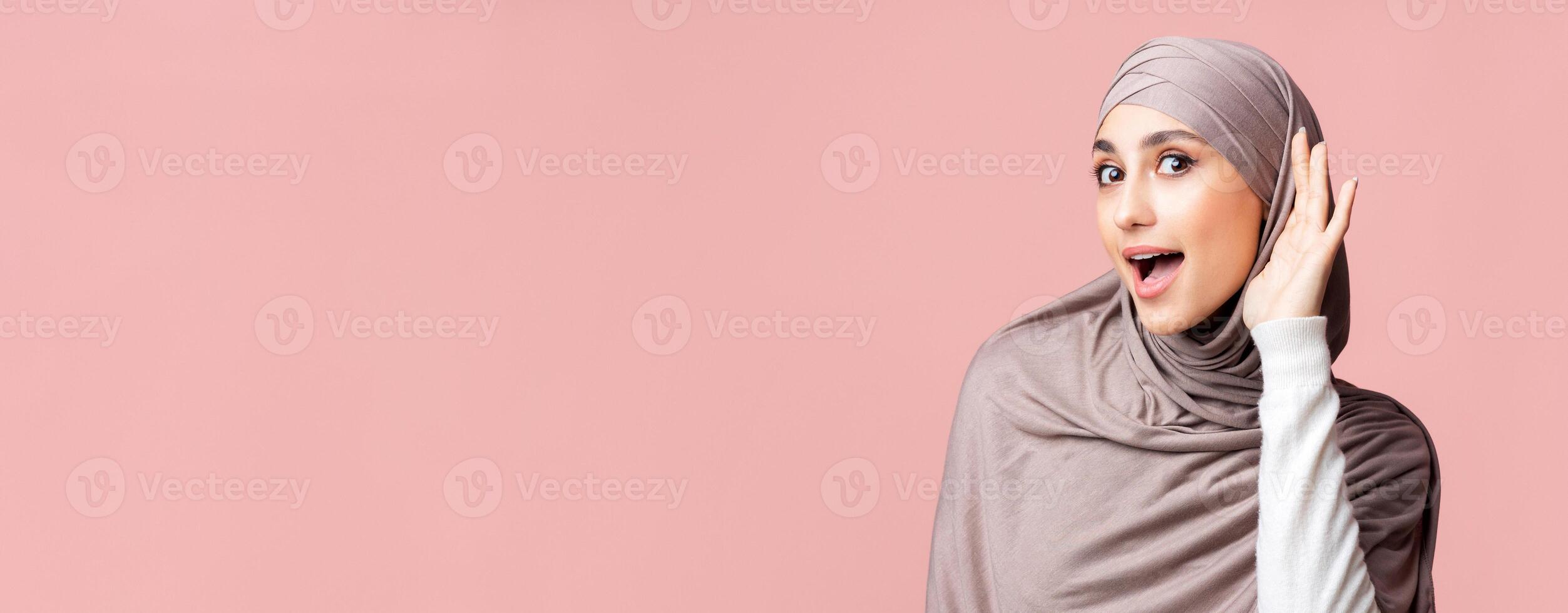 Interested arabic woman in hijab trying to hear information, holding hand near ear over pink studio background, panorama with empty space photo