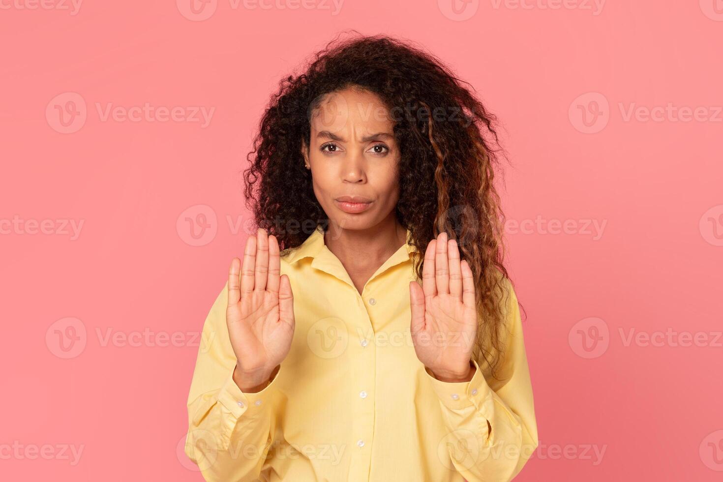 African American woman with curly hair is standing in front of a pink background, with both of her hands outstretched, palms forward, as if to say stop. photo