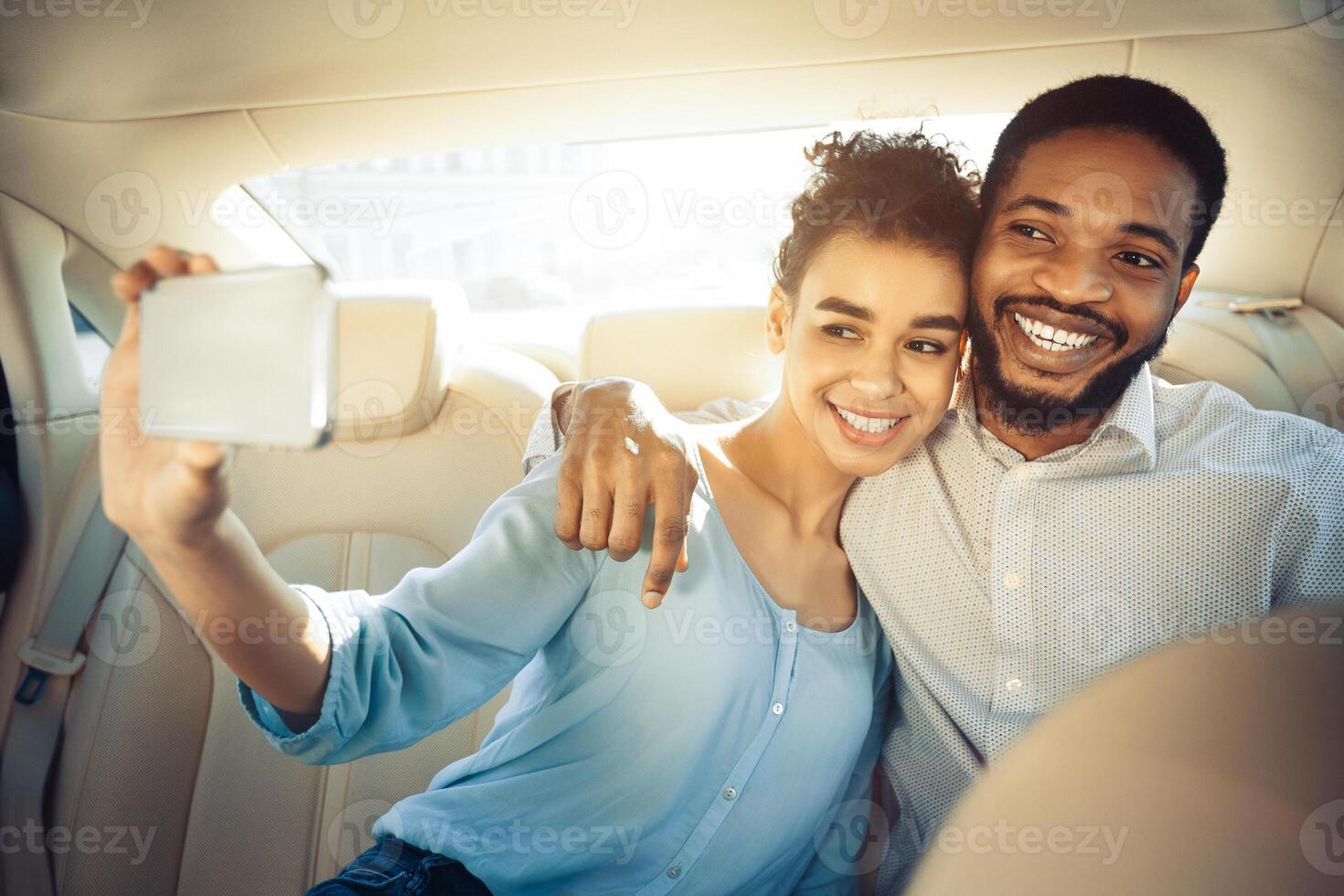African American happy couple takes a selfie in the backseat of a car. The woman is holding a phone and smiling, while the man is smiling and has his arm around her photo