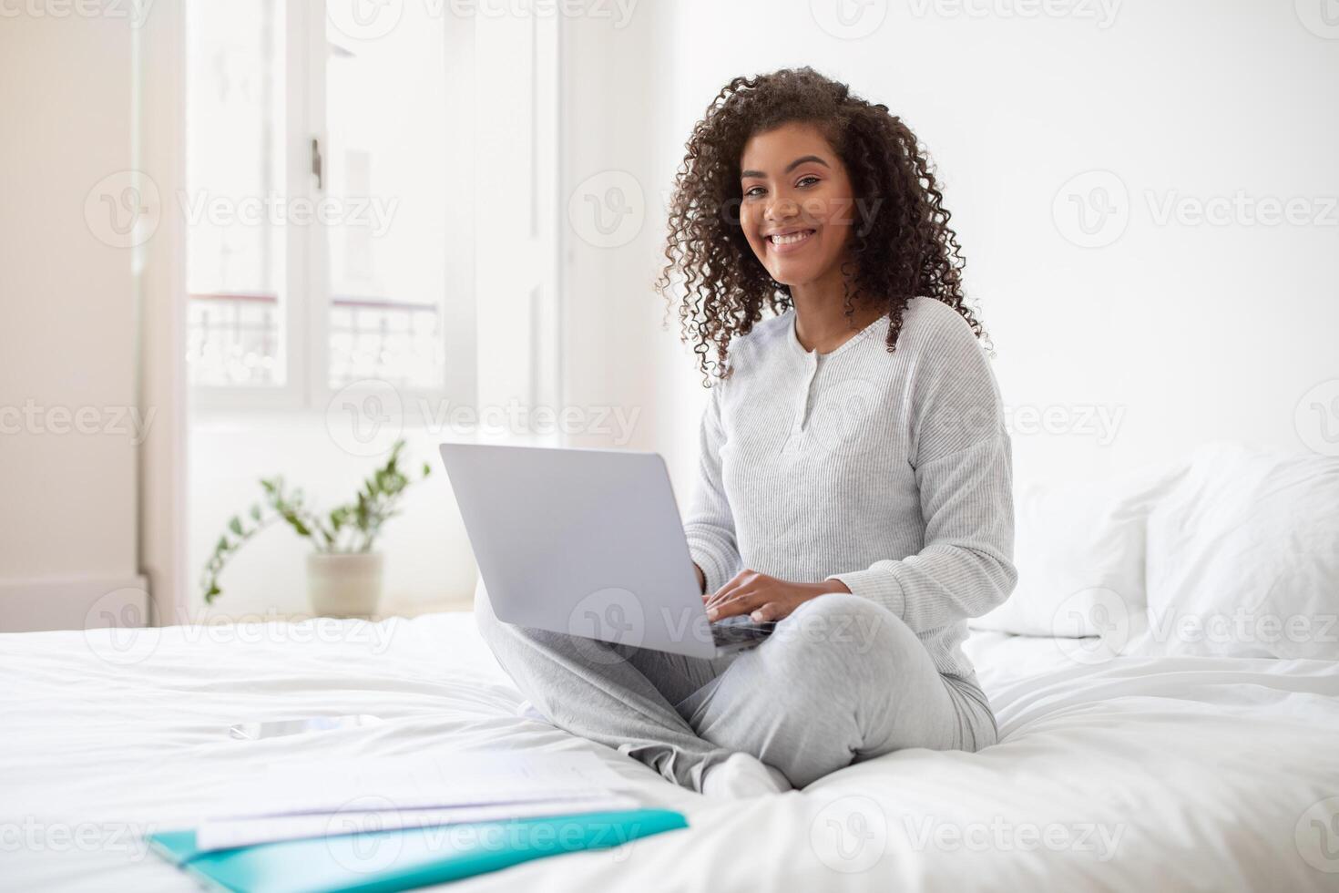 A Hispanic woman sits on a bed, focused on using a laptop computer. She types on the keyboard, with the screen displaying data. The room is well-lit, with a simple and clean decor. photo