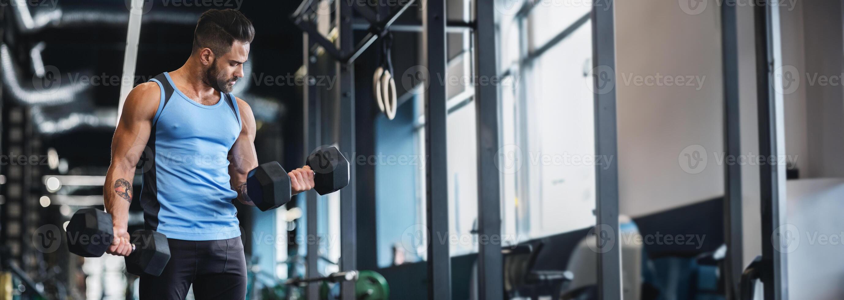 A man in a blue tank top is performing bicep curls with dumbbells in a well-lit gym, copy space photo