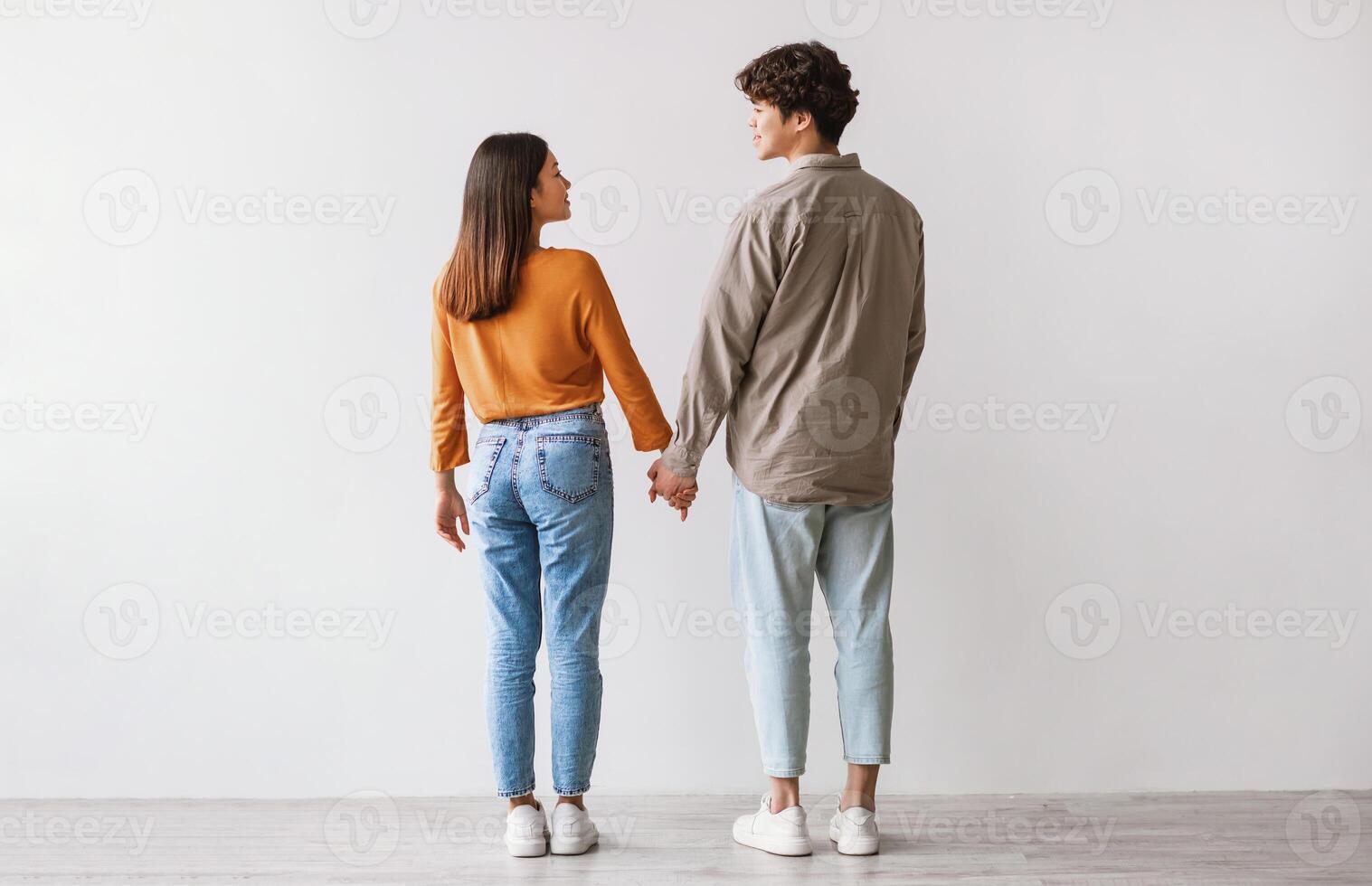 Full length of young Asian man and woman standing with their backs to camera, looking at each other and holding hands against white studio wall. Millennial couple expressing affection photo