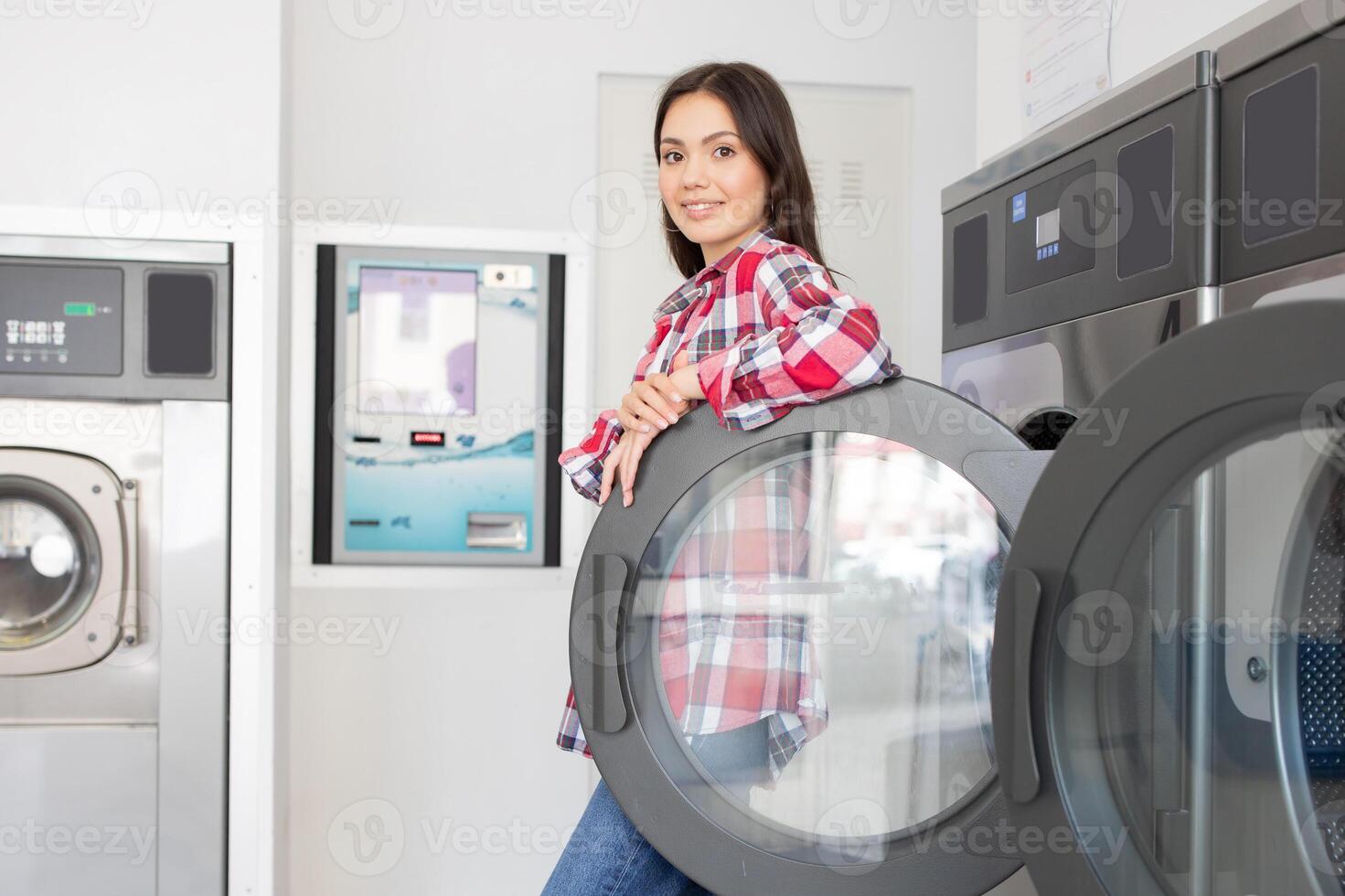 A young woman casually leans against a dryer in a contemporary laundromat, showcasing a friendly smile as she enjoys the experience of doing laundry during daytime. photo