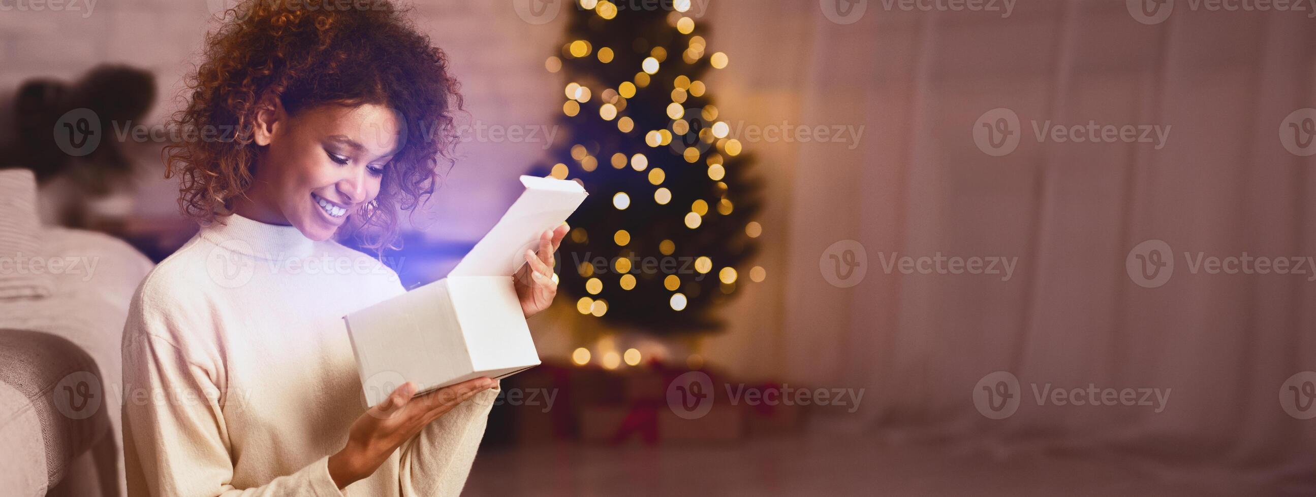 Happy black woman open xmas gift box on Christmas eve, enjoying cozy evening photo