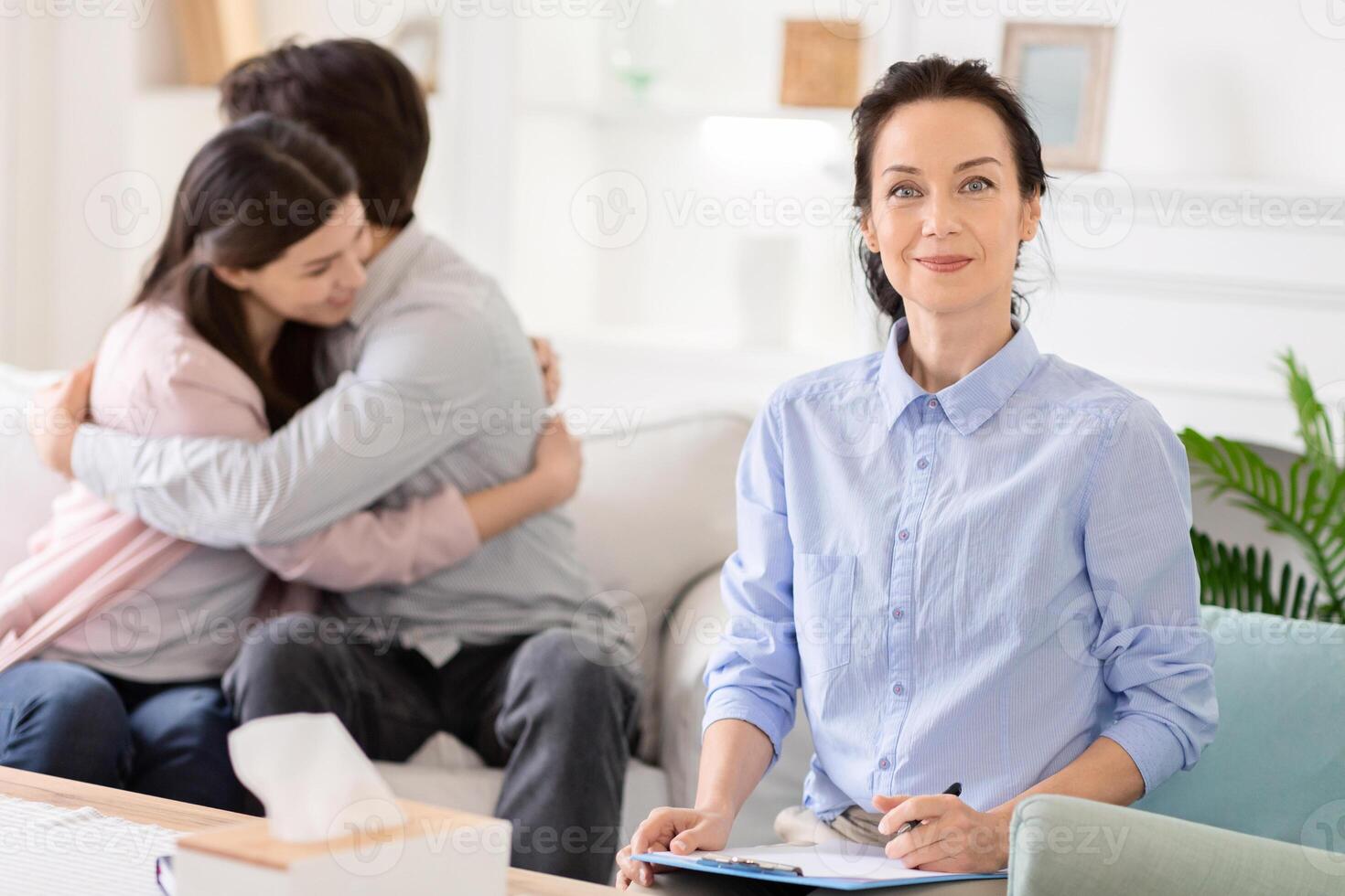 A couple hugs each other during a morning counseling session while a therapist takes notes and offers a confident smile in a bright, well-lit room. photo
