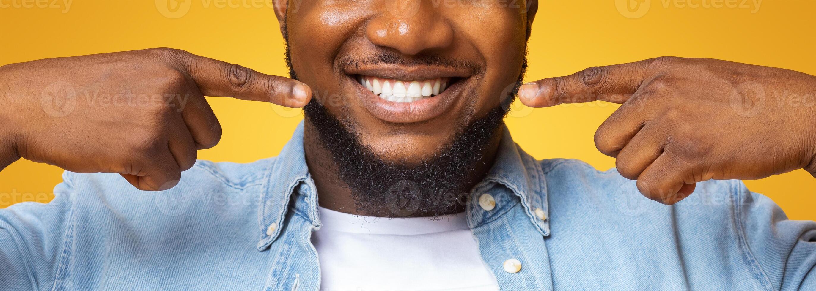 Dental health. Closeup of handsome black guy smiling wide and pointing at his perfect teeth, yellow background with copy space photo