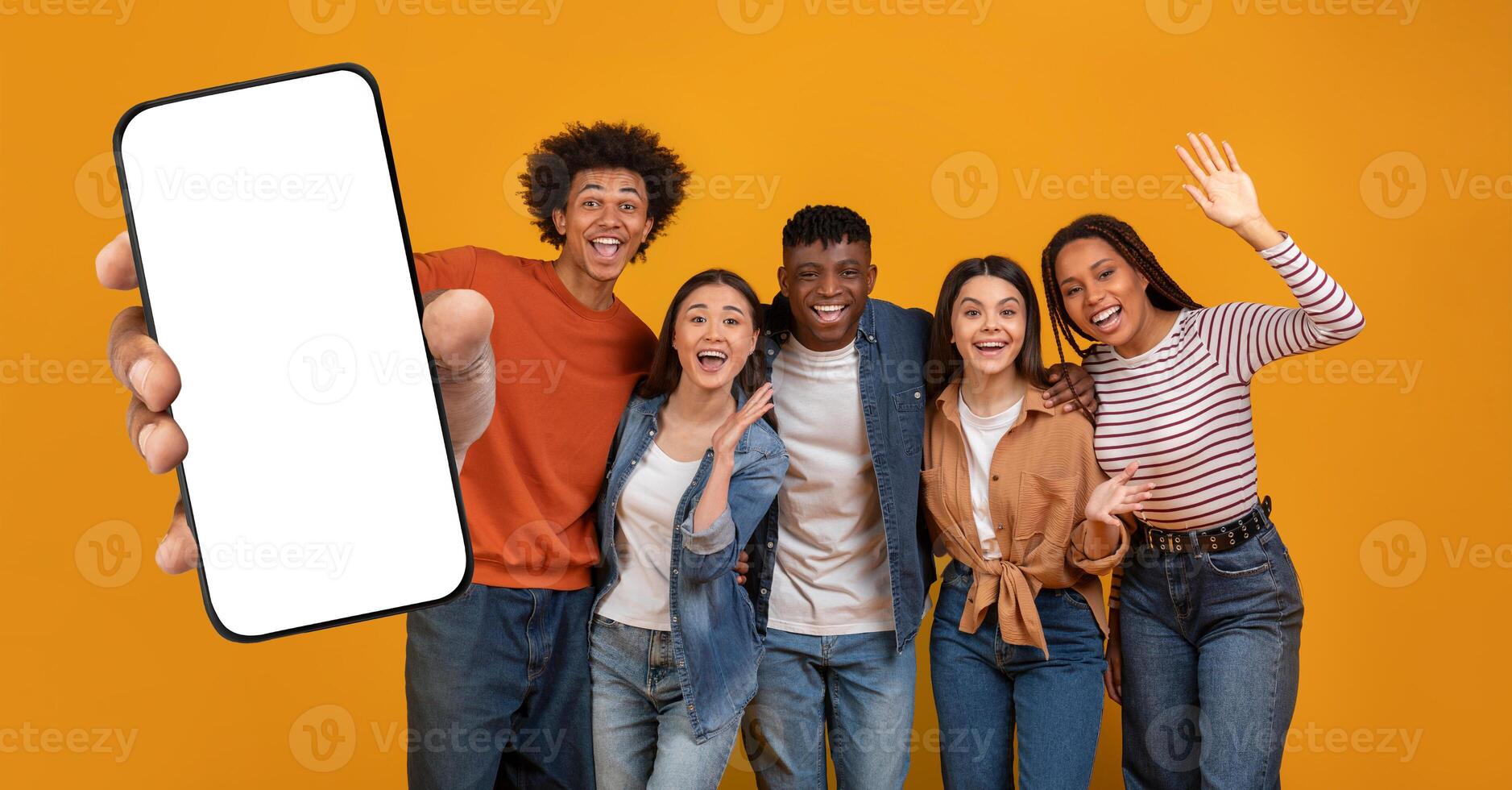 Five multiethnic friends stand together, smiling and posing enthusiastically, with one holding up a blank smartphone. The cheerful atmosphere is highlighted by a vibrant yellow backdrop. photo