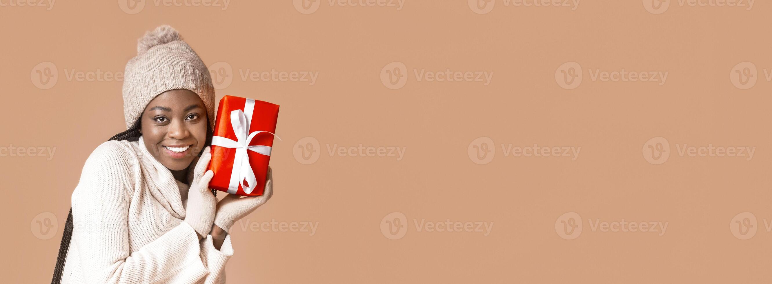 Happy black woman in winter clothes with christmas gift over beige studio background, copy space photo