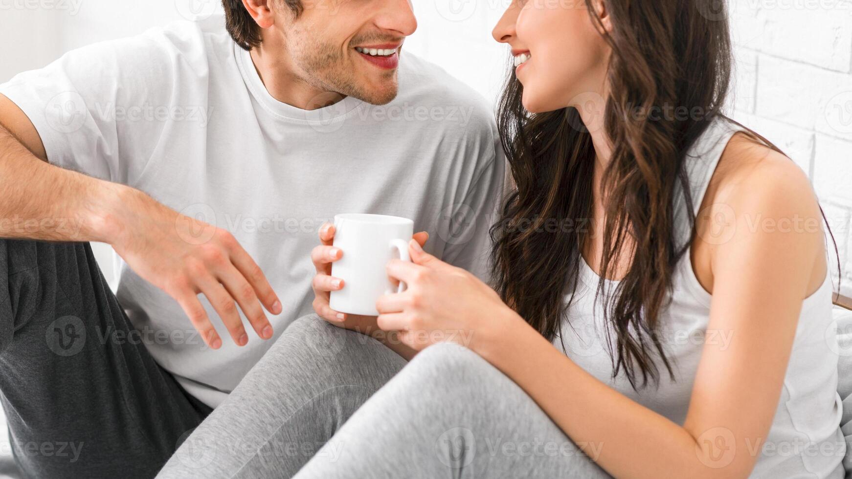 A smiling man and woman are sitting on a bed together, enjoying their coffee in the morning. They are looking at each other, and the woman is holding her coffee mug in her hands, cropped photo