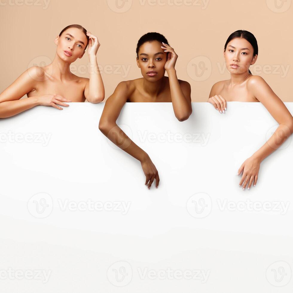 Three Multiracial Girls Posing Standing Naked Behind White Poster With Blank Space For Text On Gray Background. Studio Shot photo
