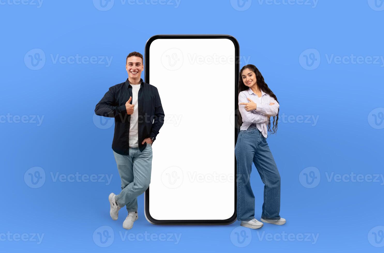 Smiling multiethnic young man and woman standing beside a large smartphone with blank screen mockup, isolated on blue background. photo