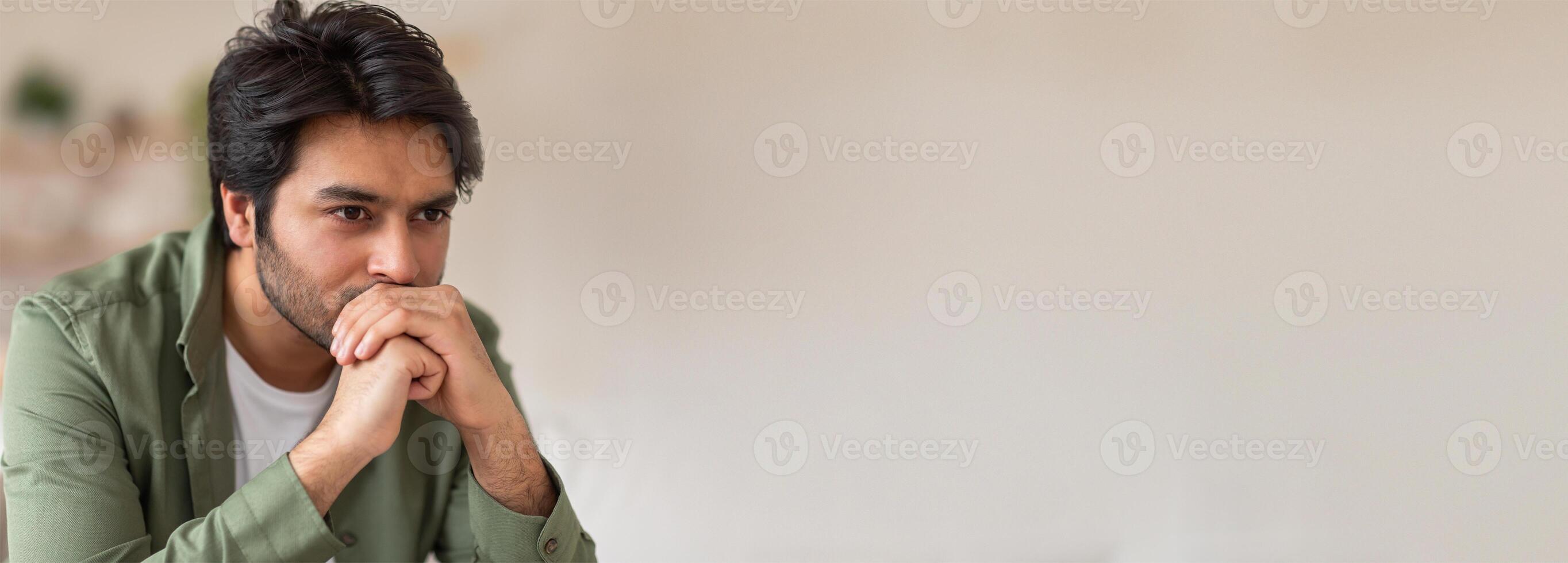 A young Indian man sits in a well-lit room, deep in thought with his hands clasped, indicating contemplation or problem-solving. Natural light enhances the serene atmosphere. photo