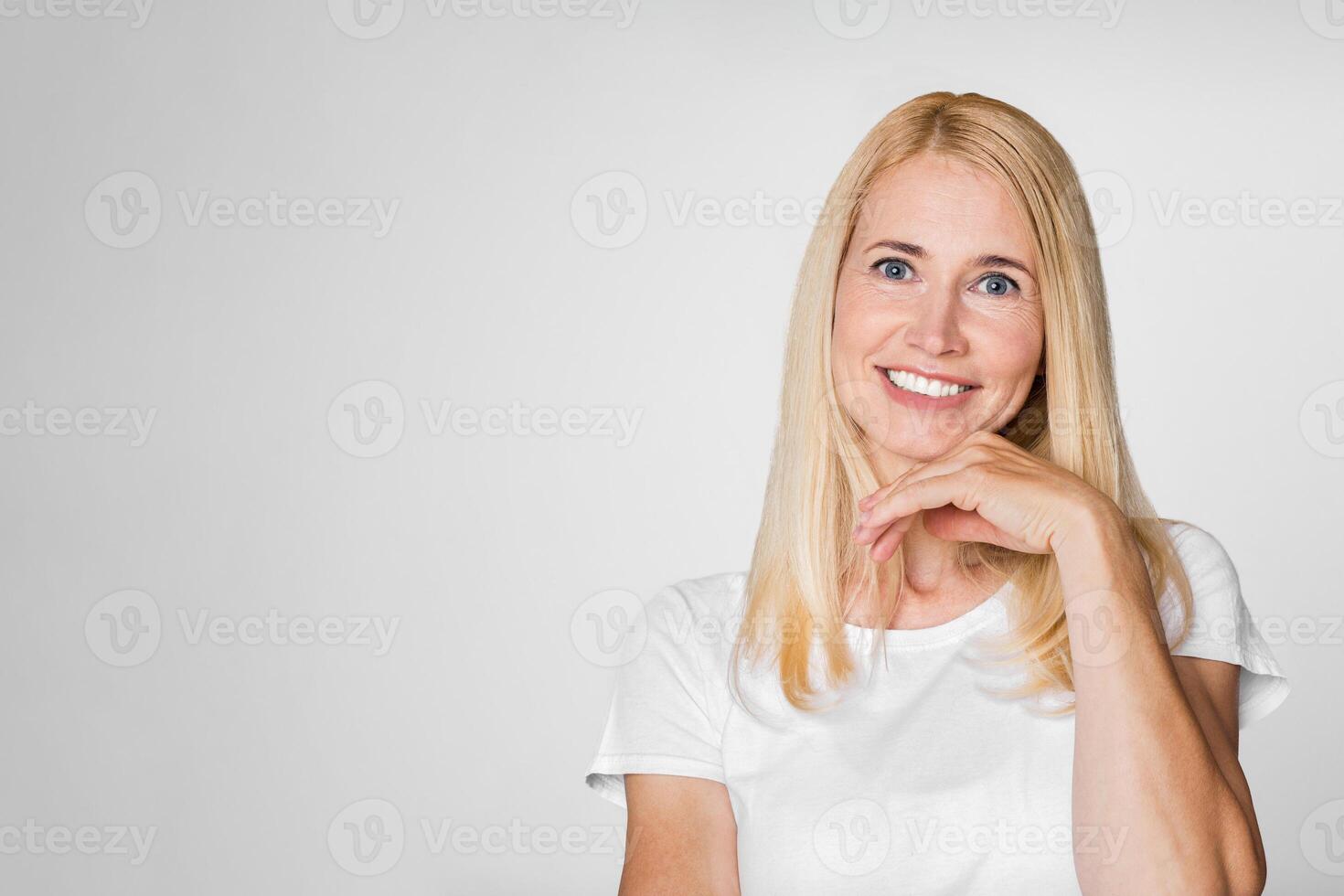 A portrait of a woman with long blonde hair, wearing a white t-shirt, smiling and looking at the camera. Her right hand is resting on her chin. The background is a plain white wall. photo