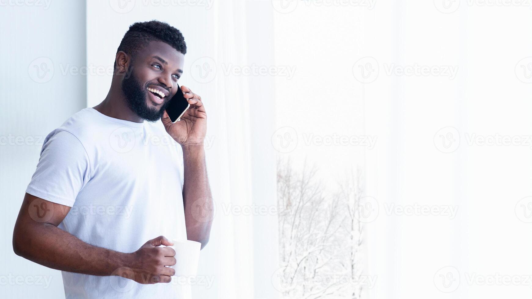 African American man with a beard smiles while talking on his phone. He is holding a cup in his hand and is standing in front of a window. The man is wearing a white t-shirt, copy space photo