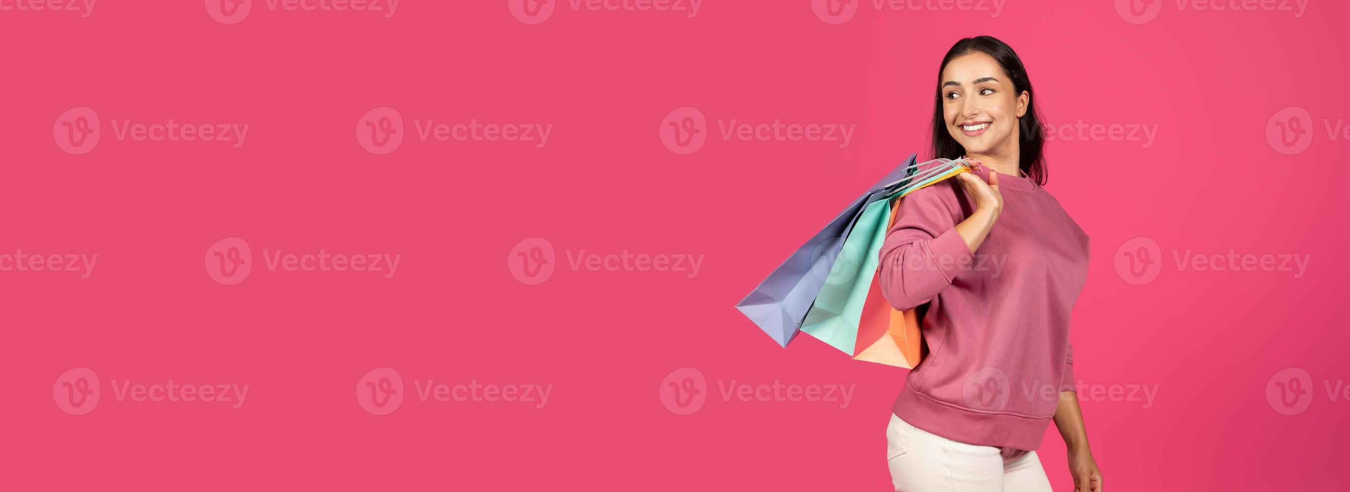 A young woman dressed in casual attire beams with joy as she carries vibrant shopping bags. The bold pink backdrop adds to the cheerful atmosphere, reflecting her excitement, copy space photo