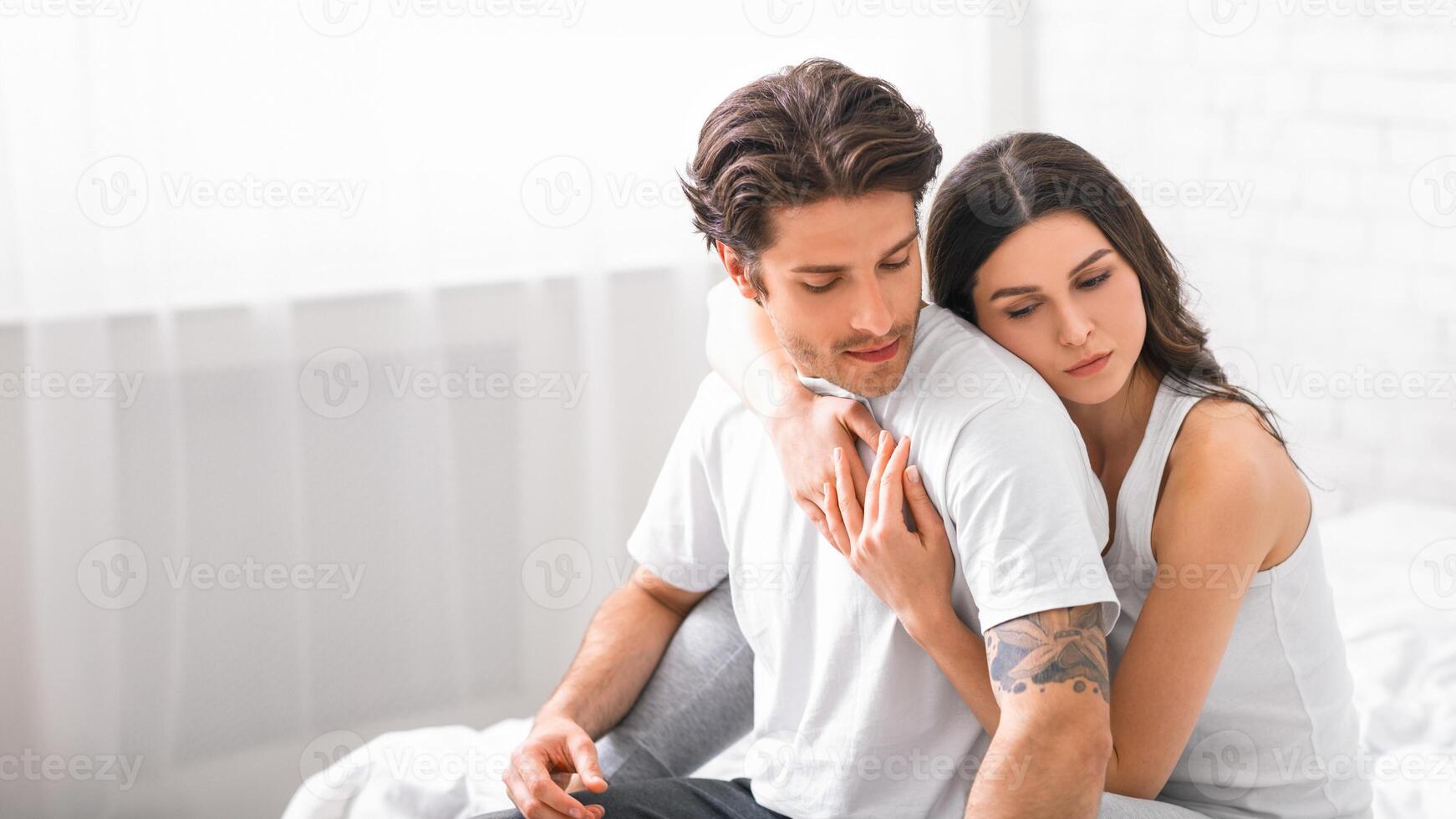 A young man and woman are sitting on a bed, the woman has her arms wrapped around the mans shoulders as she leans her head on his shoulder. Both are wearing white shirts, copy space photo