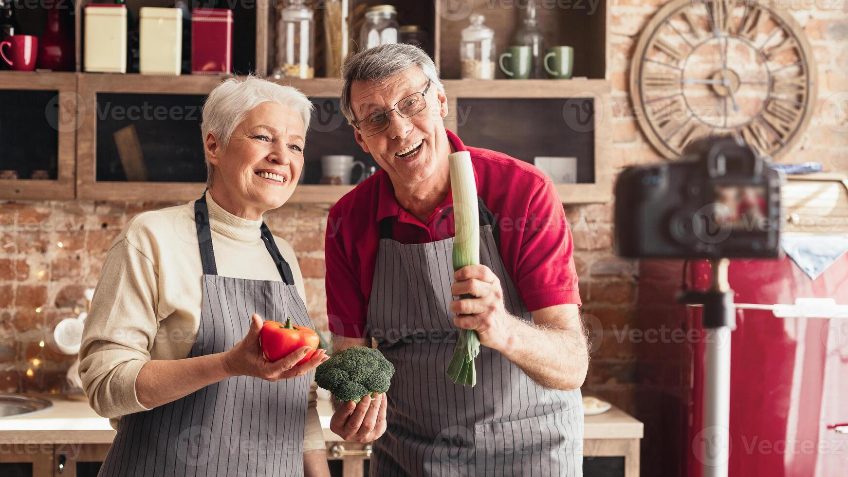 A senior couple is filming a cooking show in their home kitchen. The woman is holding a red cabbage while the man is gesturing with his hand and smiling at the camera. photo