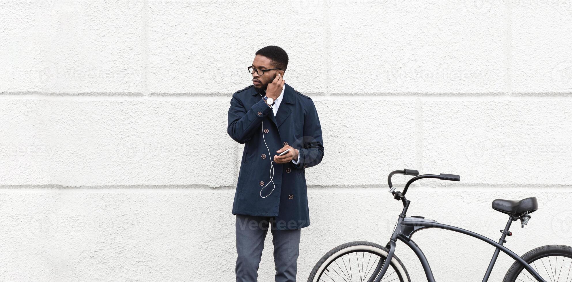 An executive black guy is cycling in an urban city environment on his way to work, standing over white wall, using earphones, listening to music, copy space photo