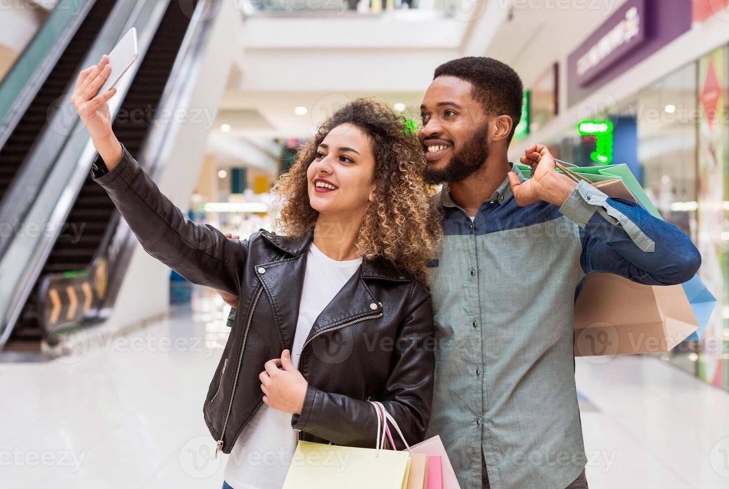 African American man and woman are seen using a smartphone to take a selfie inside a bustling shopping mall. They are smiling and posing together, capturing a moment of togetherness photo