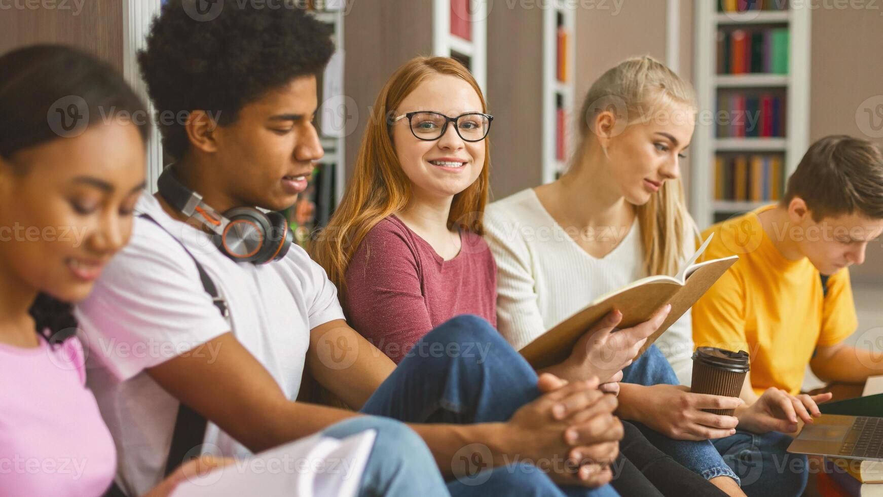 Concentrated international classmates sitting on wooden floor at library in campus and preparing for exams with books and laptops photo