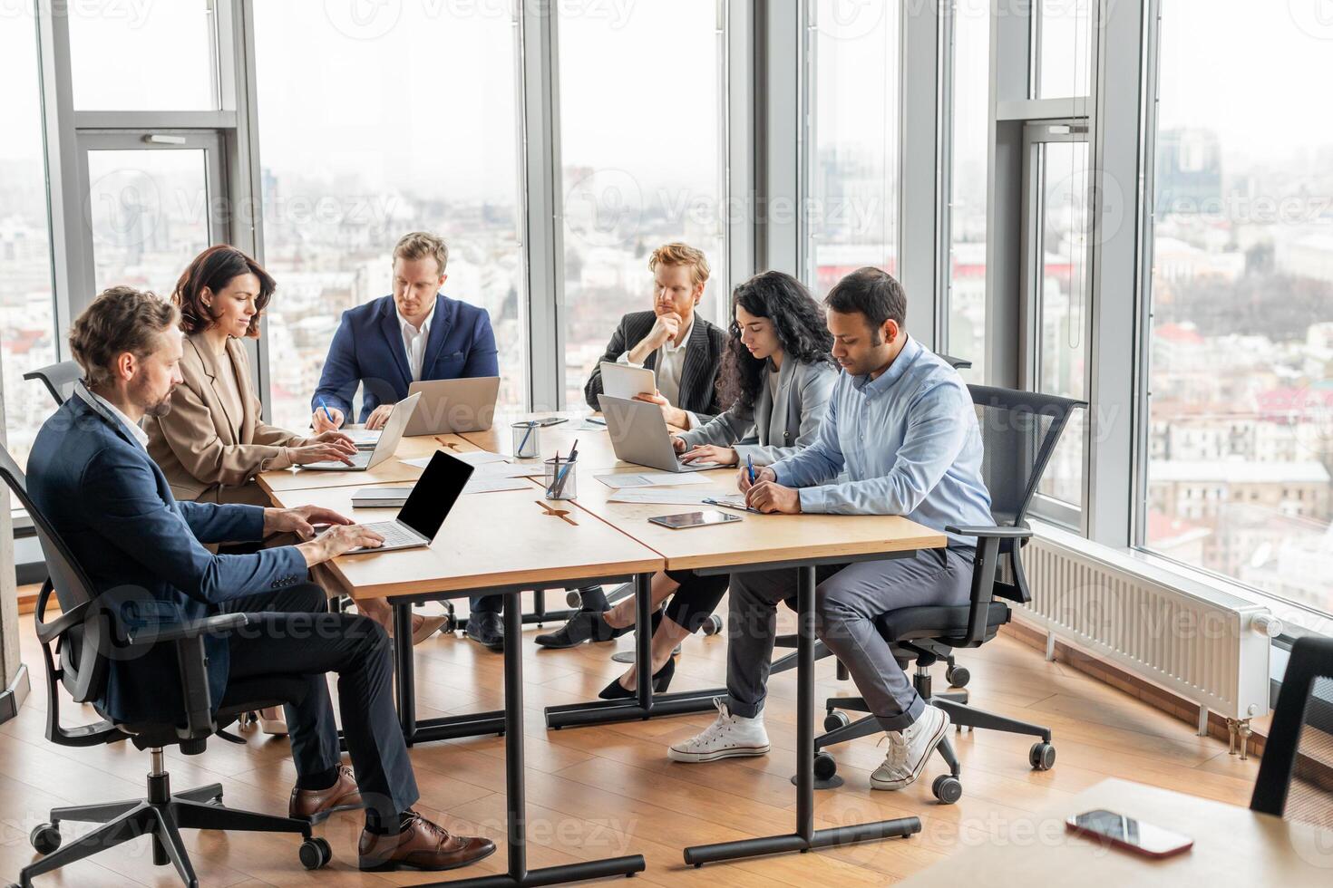 A group of business professionals work on their laptops at a large wooden table in a modern office. The office is filled with natural light from large windows overlooking a city. photo