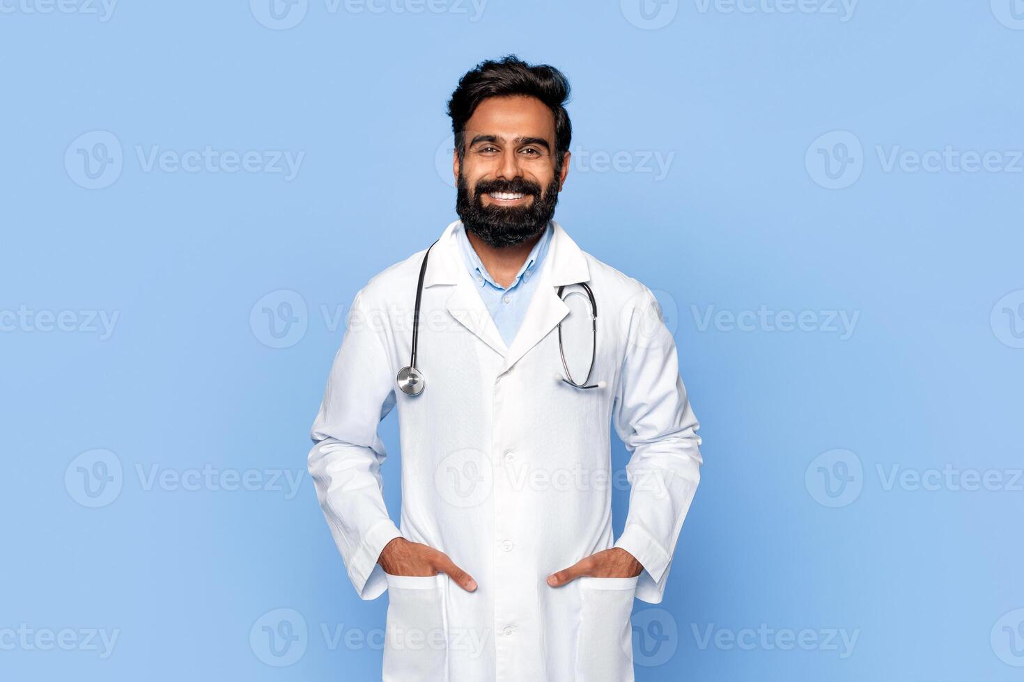 Indian man with a beard and dark hair is wearing a white doctor's coat with a stethoscope around his neck. He is smiling at the camera and has both hands in his pockets. photo