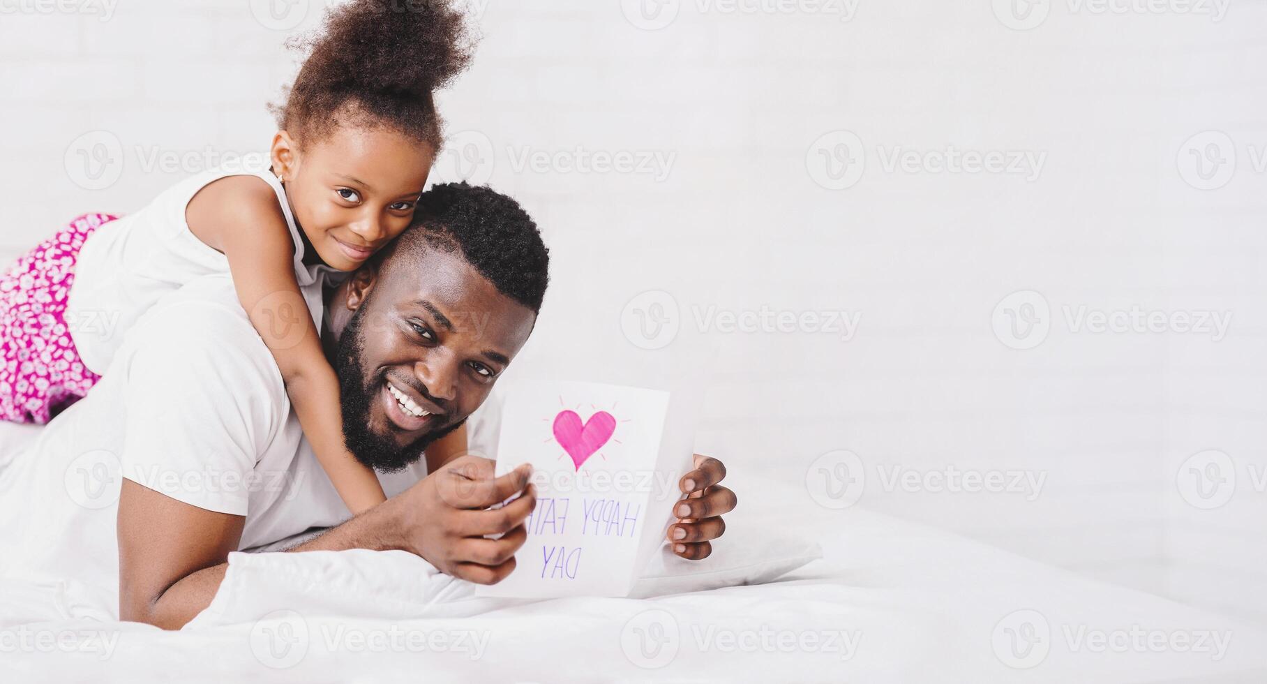 Black father is lying in bed holding a card that has a pink heart and handwritten words on it, smiling at the camera. His daughter is sitting on his back, her arms wrapped around his neck photo