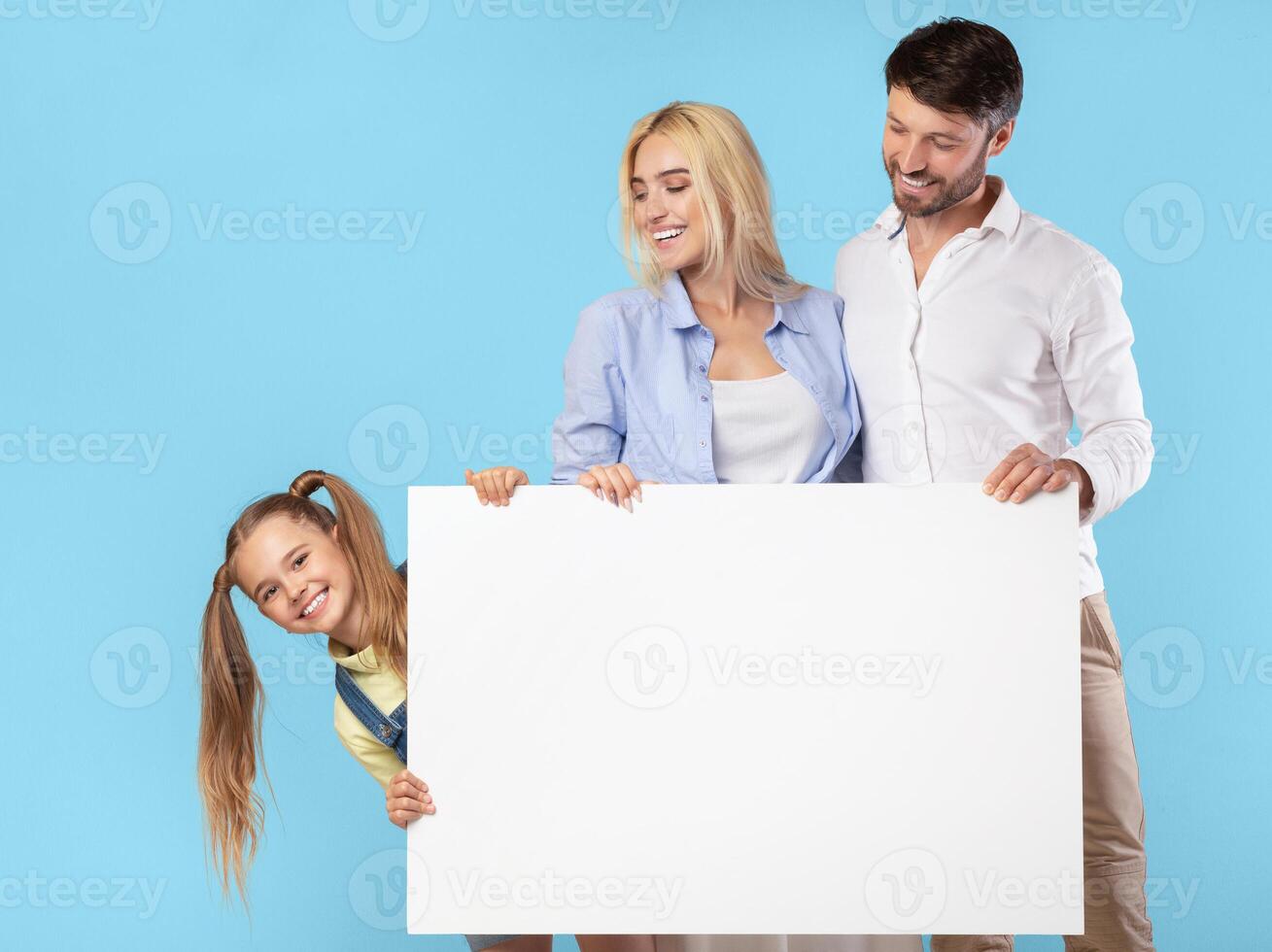 A family of three stands in front of a blue background, holding a large blank white sign. The parents, a man and a woman, smile and look at the sign. Their young daughter peeks around the side photo