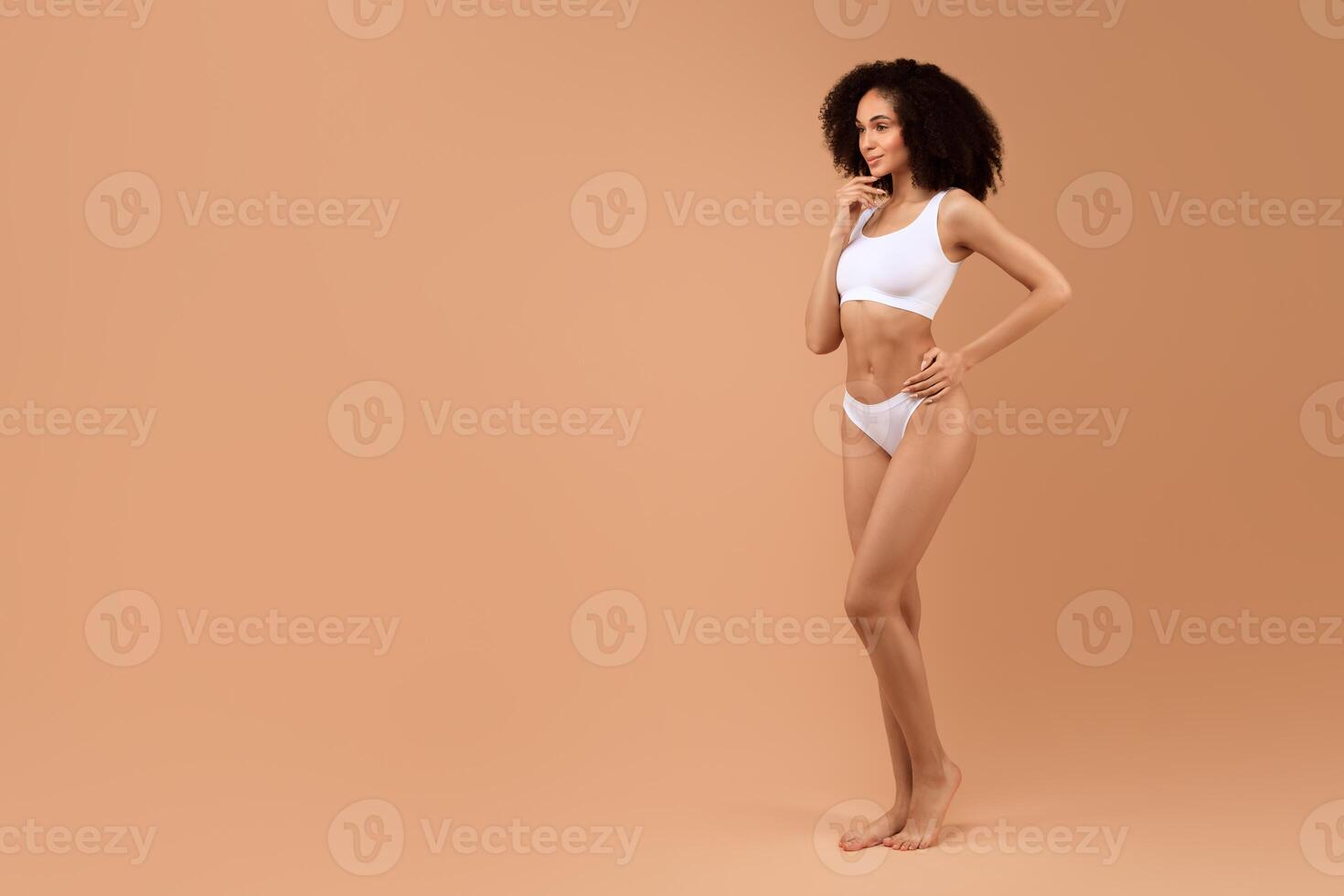 Black woman in white lingerie stands with one hand on her hip and the other on her chin. She is looking at copy space and has a contemplative expression. The background is a simple beige color. photo