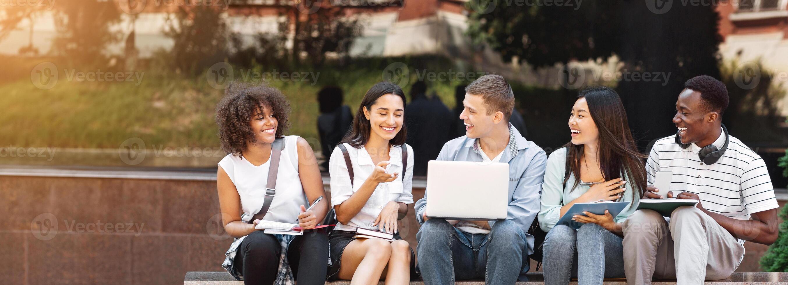 Campus Life. Cheerful college students resting outdoors between classes, using laptop, chatting and laughing, free space photo